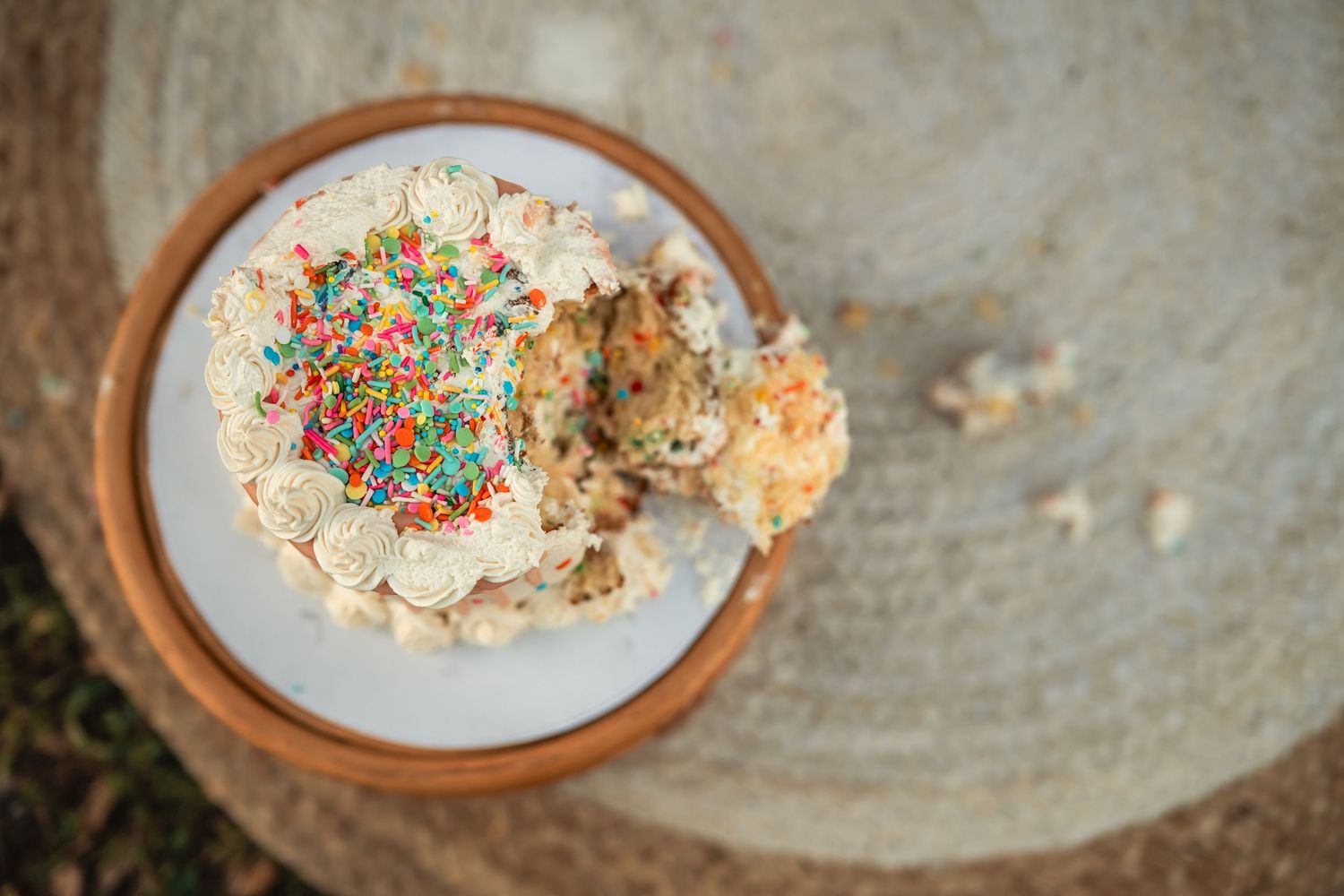 Top view of a partially eaten birthday cake with colorful sprinkles on a white plate atop a wooden stump.