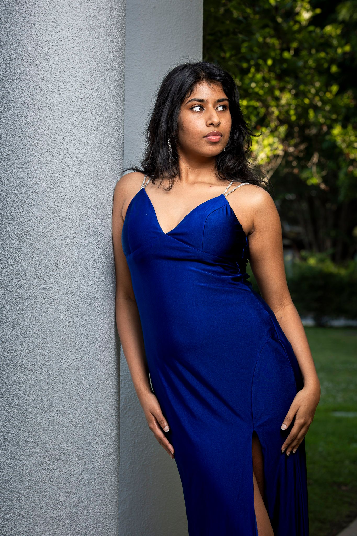 Young woman in elegant royal blue satin gown with spaghetti straps, leaning against white pillar outdoors.