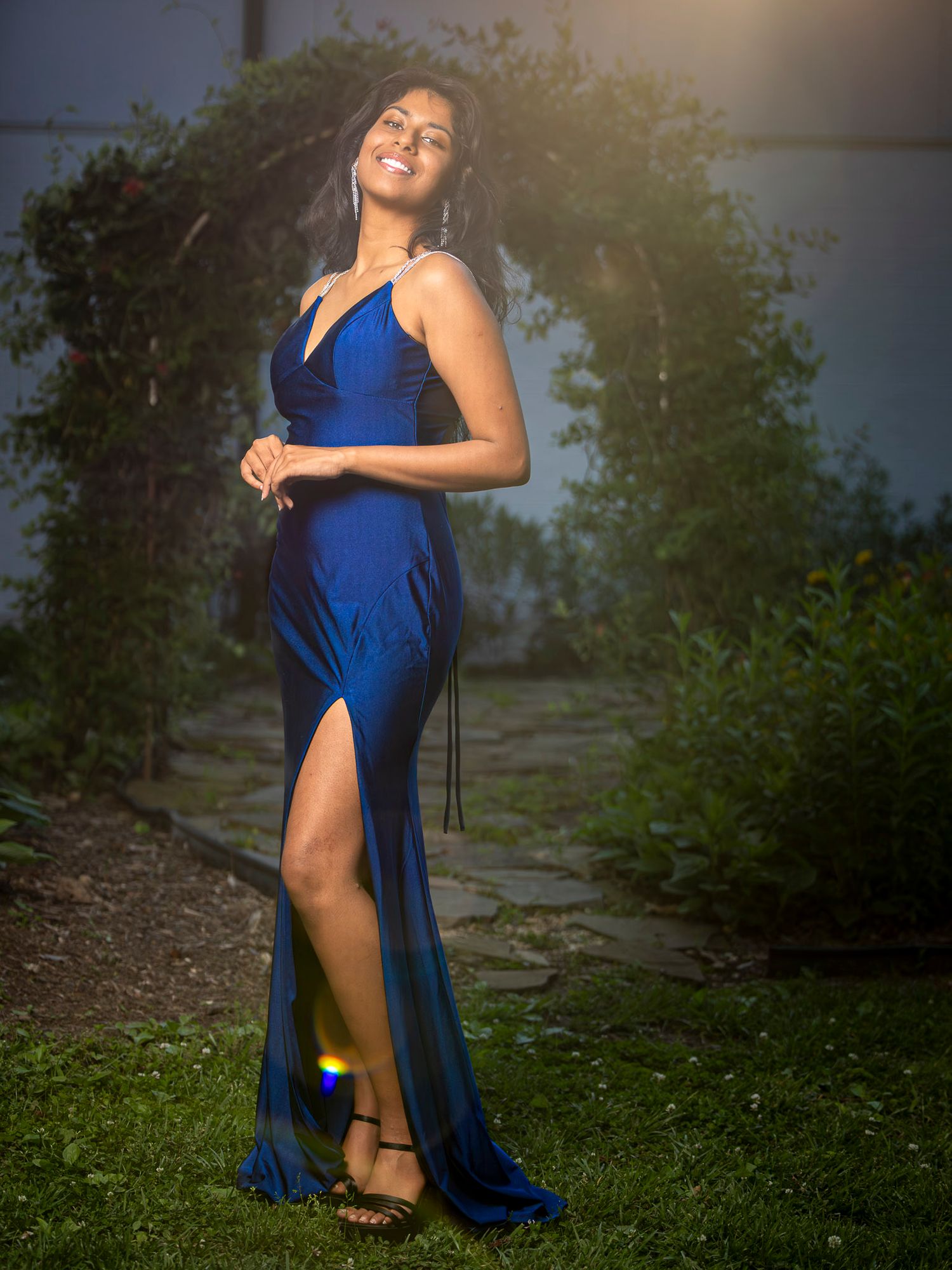 Woman in elegant royal blue satin gown with thigh-high slit poses gracefully outdoors at night.
