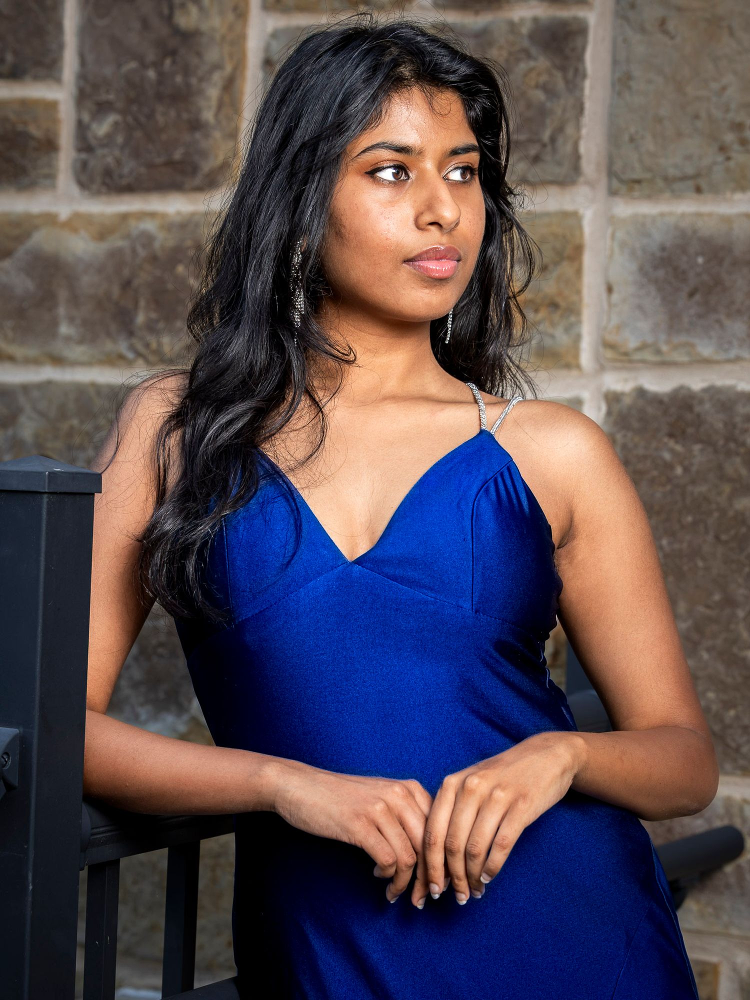 Young woman in royal blue satin slip dress poses elegantly against stone wall backdrop.