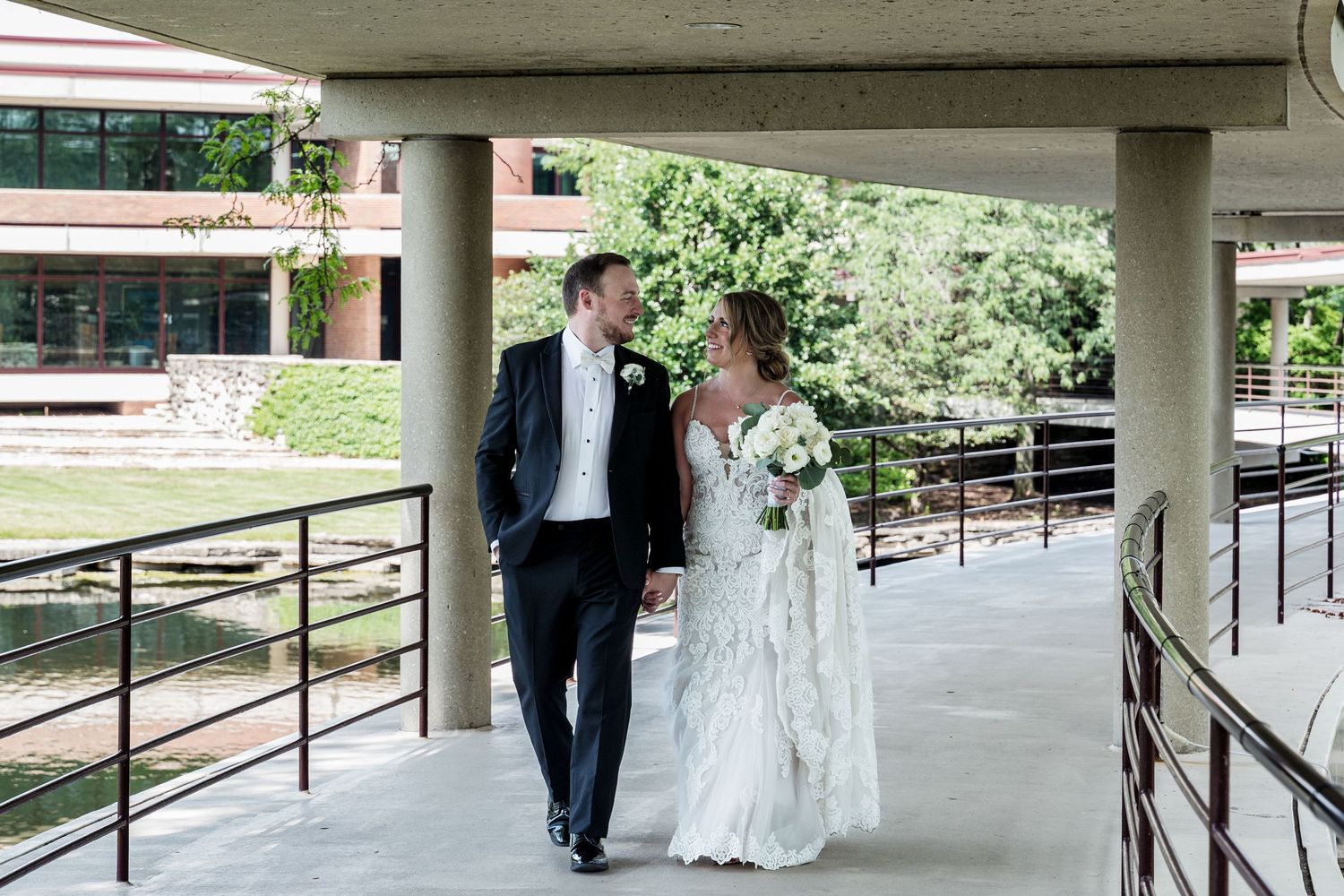 Bride and groom walking hand in hand under a covered walkway on their wedding day, surrounded by lush greenery.