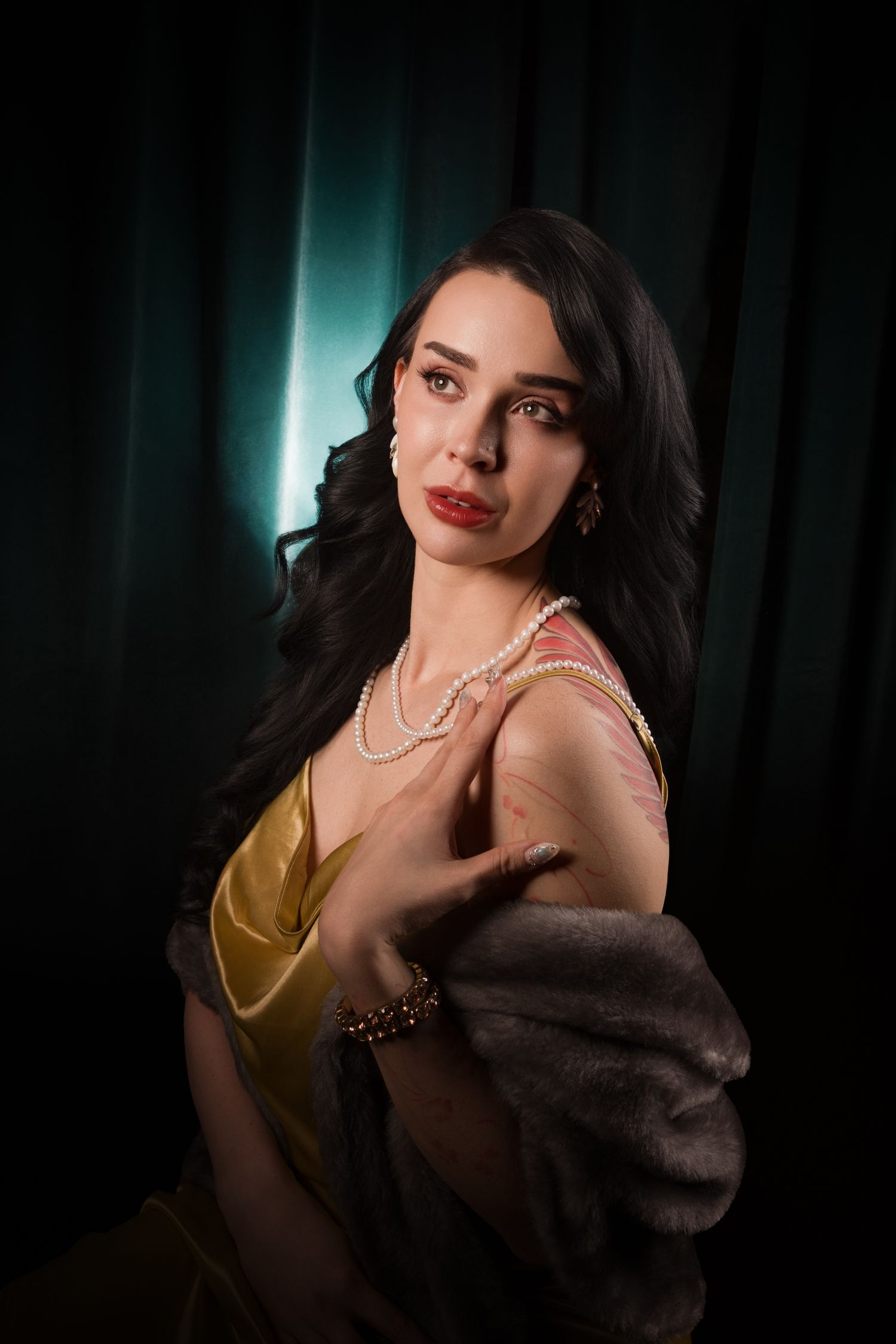 Elegant woman in gold dress with pearl necklace and fur stole poses against dark teal curtain backdrop.