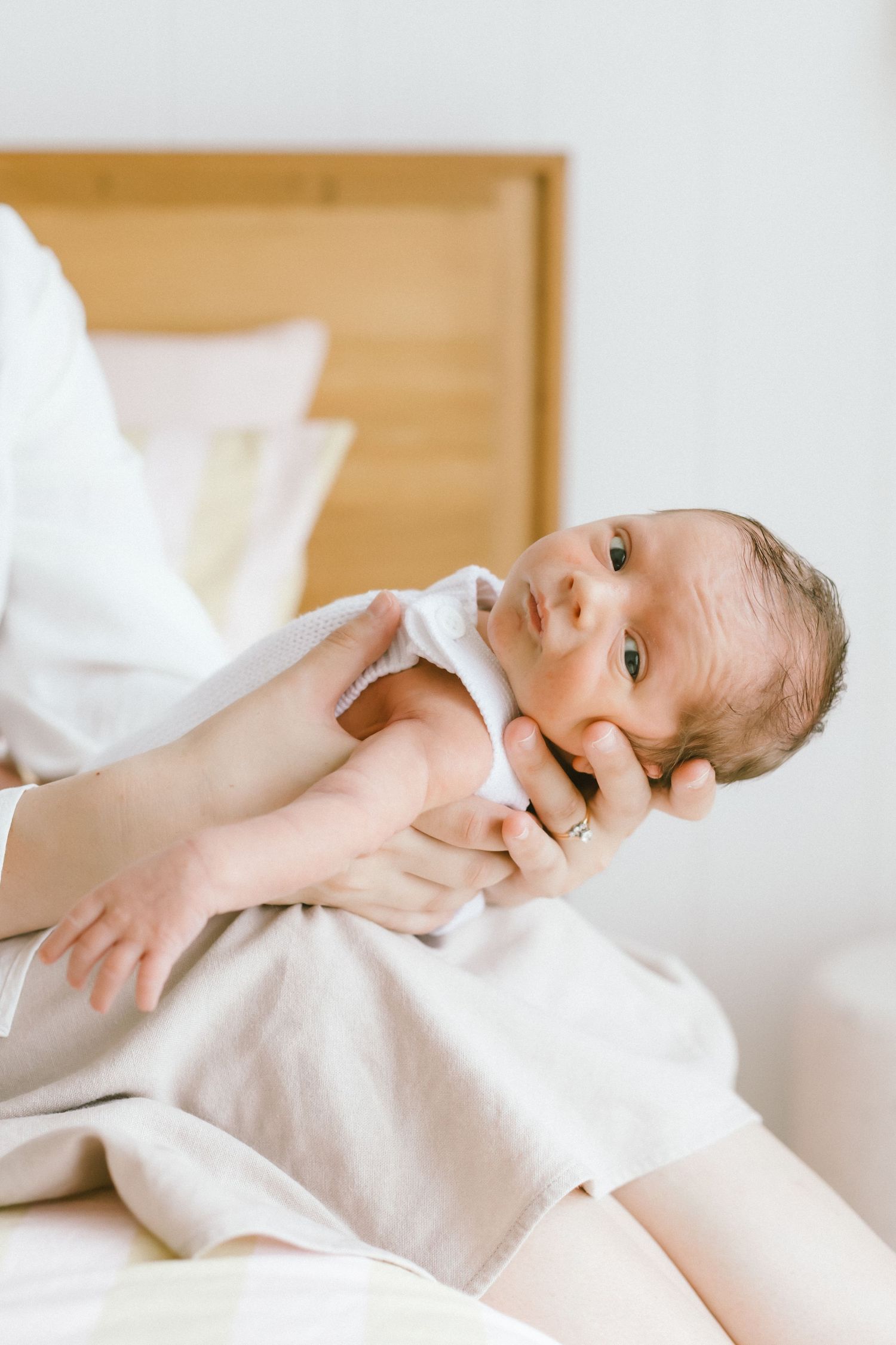 Mother gently holding newborn baby in white onesie while sitting on bed in bright, minimalist nursery.