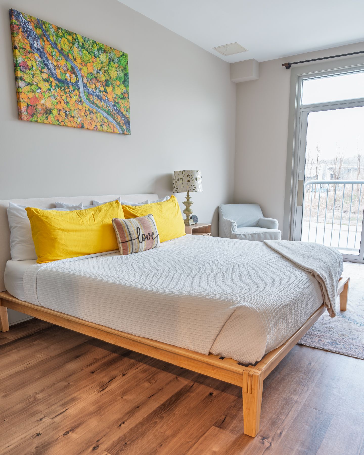 A bright bedroom with a king bed, yellow pillows, hardwood floors, and a colorful tree painting above the headboard.
