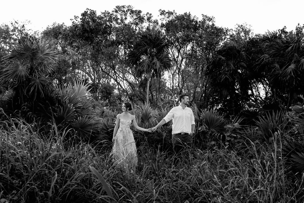 Black and white photo of an engaged couple holding hands while walking through lush tropical vegetation during their engagement session.