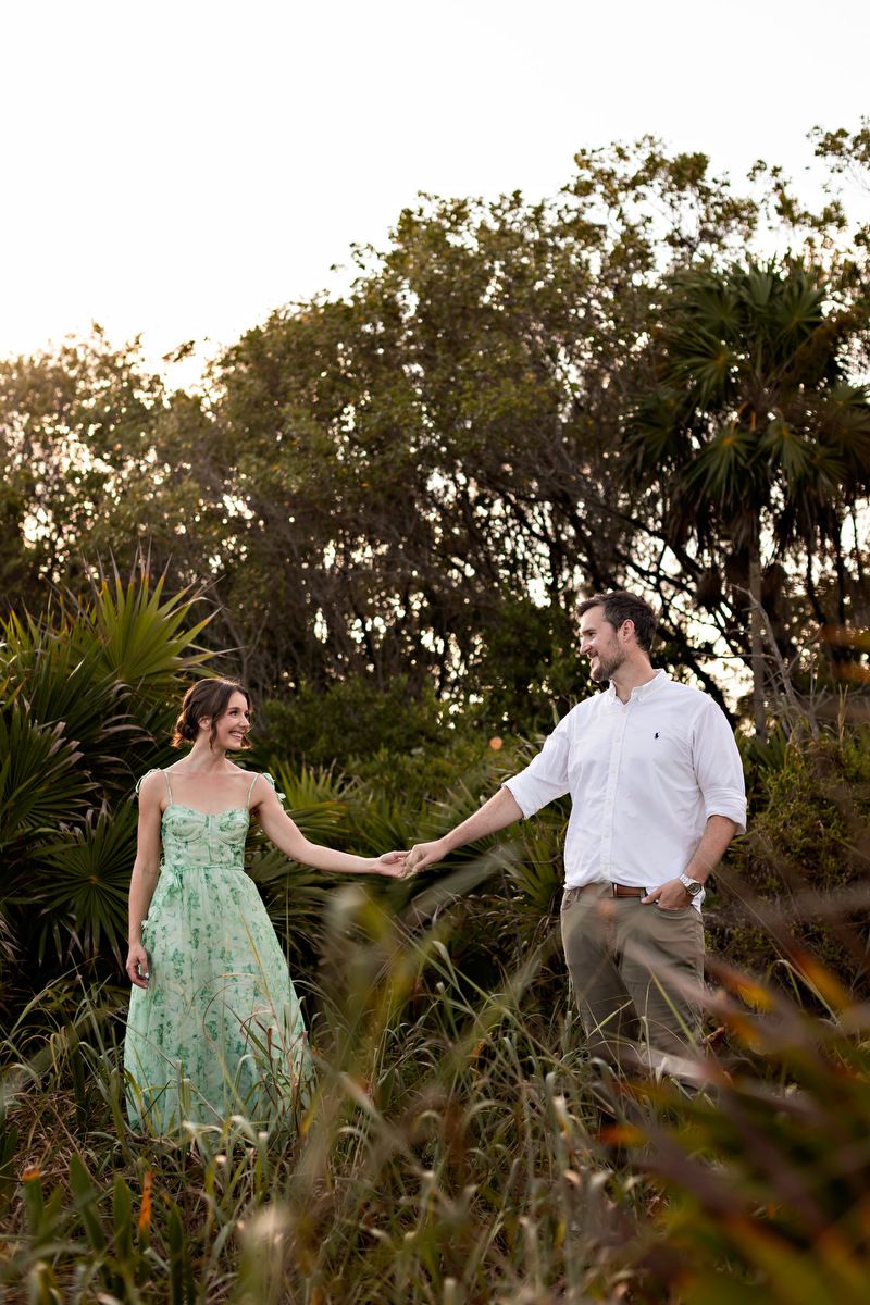 Romantic couple holding hands in lush green field at golden hour, woman in mint floral dress, man in white shirt.