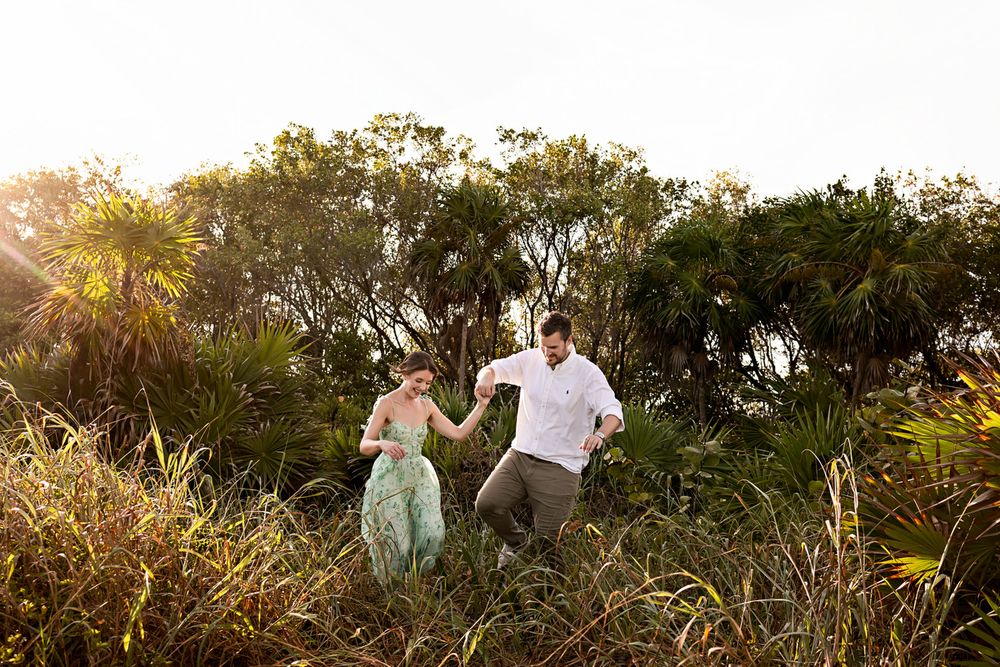 Couple holding hands dancing through tall grass and tropical foliage during golden hour sunset outdoors.