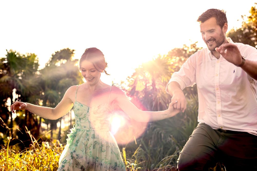 Joyful couple dancing in sunlit meadow, woman in floral dress, man in white shirt, with bright lens flare effect.