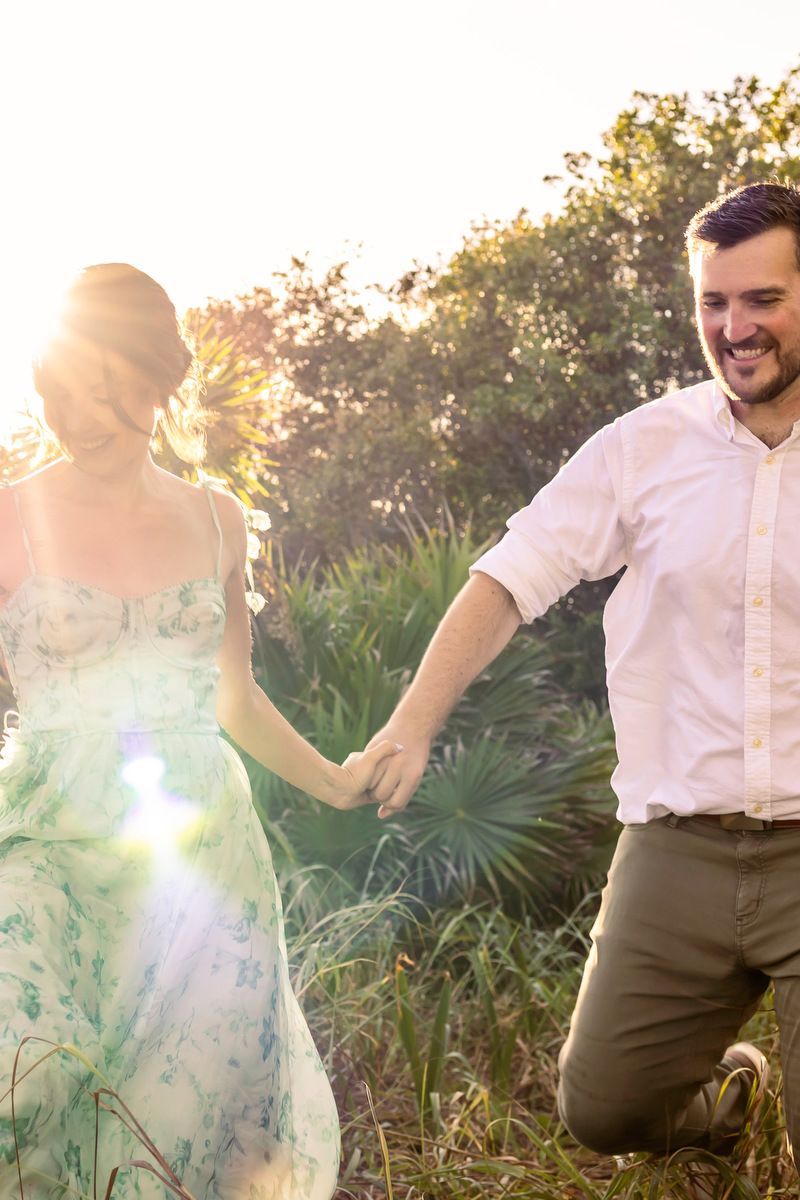 Happy couple holding hands in a sunlit outdoor setting, man smiling in white shirt, woman in floral dress.