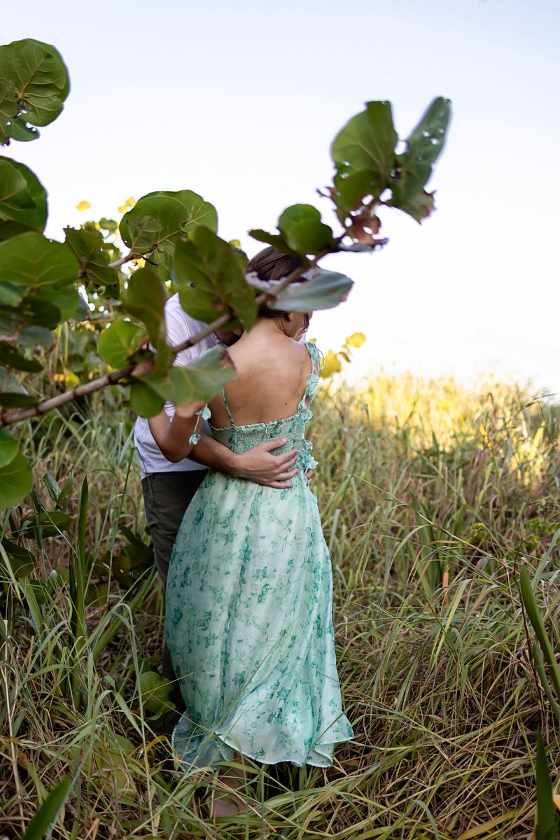 Couple embracing in tall grass field, woman wearing elegant backless green floral dress, surrounded by lush tropical foliage.