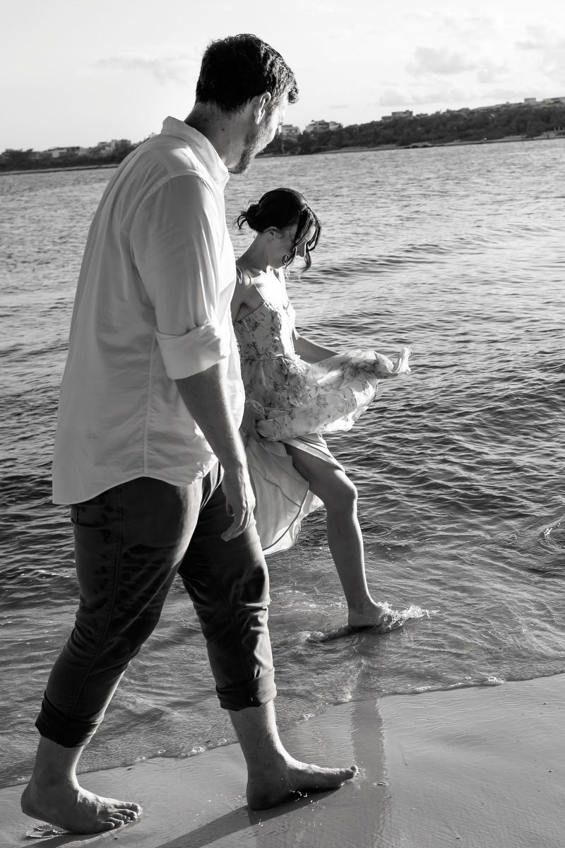 Black and white photo of a couple walking barefoot in shallow ocean waves at the beach.