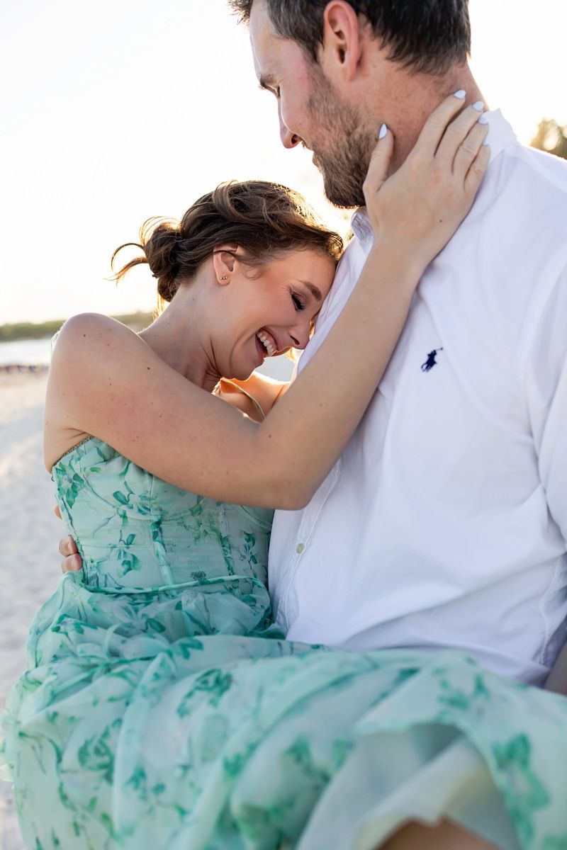 Couple embracing on a beach, woman in green floral dress laughing joyfully against man in white shirt.