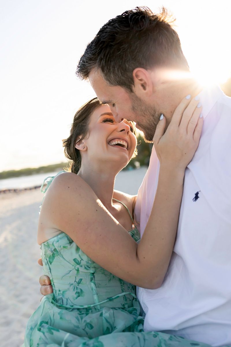 Couple laughing and embracing on a sunny beach, woman in teal floral dress, golden backlit sunset.