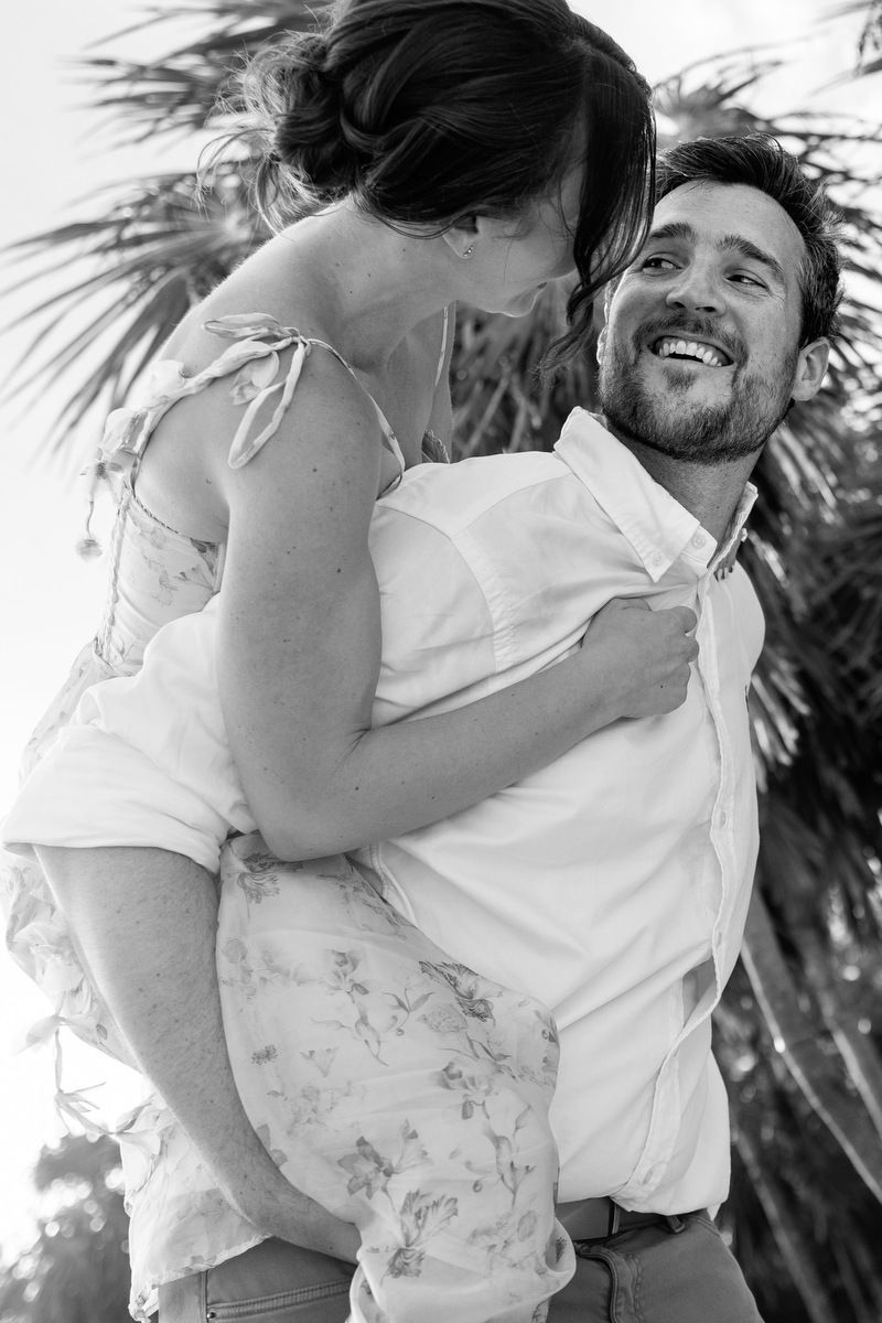 Black and white photo of a laughing man lifting a woman in a floral dress near palm trees.