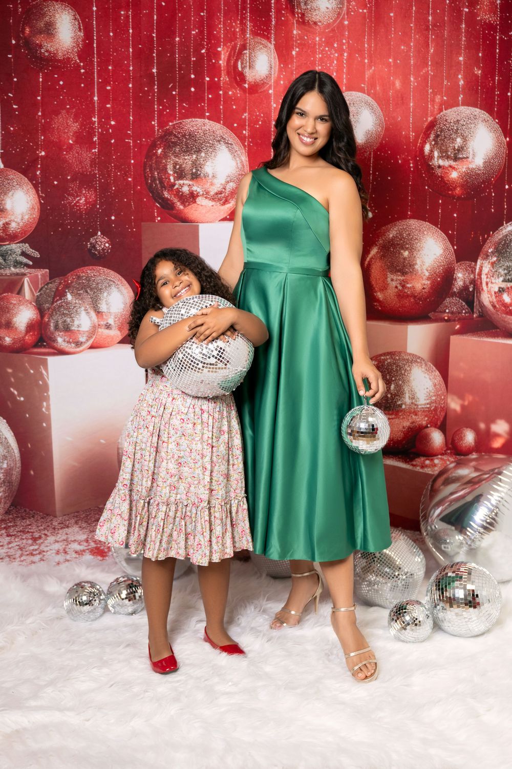 Woman in green satin dress and girl in floral dress pose with disco balls in festive Christmas setting.