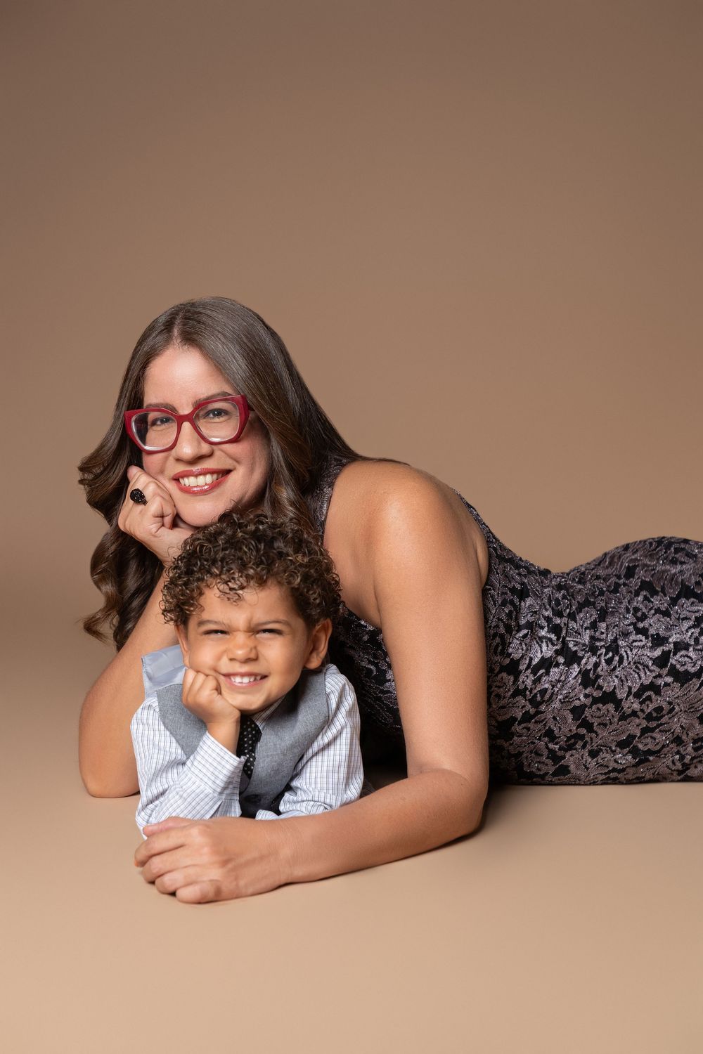 A smiling woman in glasses and a boy in a tie pose together lying on the floor against a beige background.
