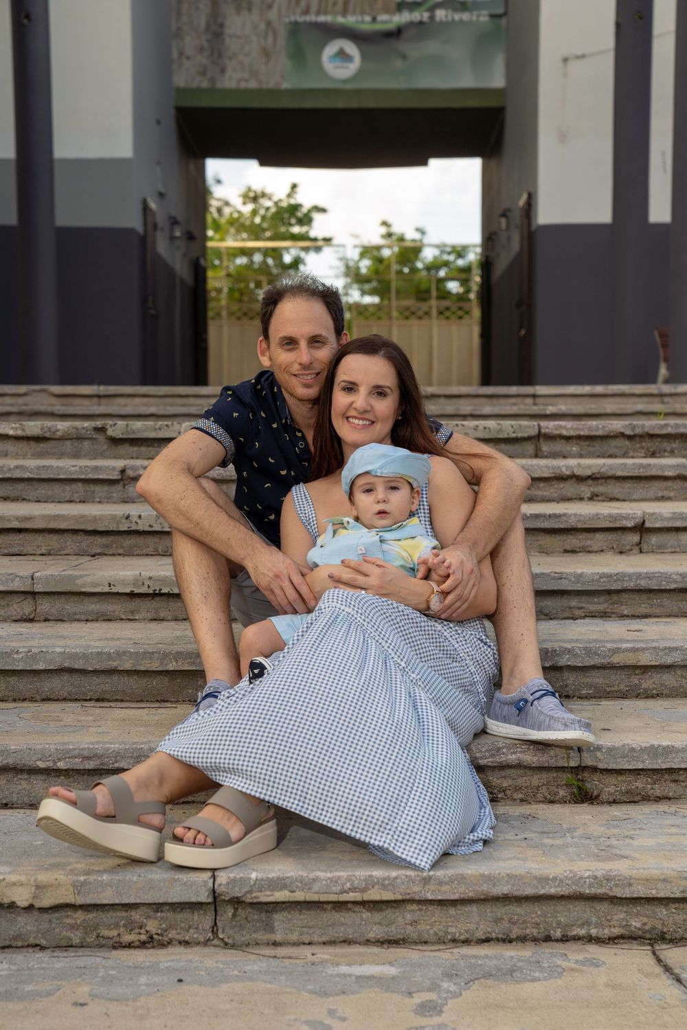 Happy family with baby sitting on stone steps, smiling couple holding infant in blue outfit outdoors.