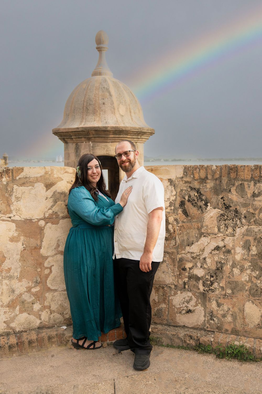 Couple posing by stone fort wall with rainbow in cloudy sky, woman in teal dress, man in white shirt.