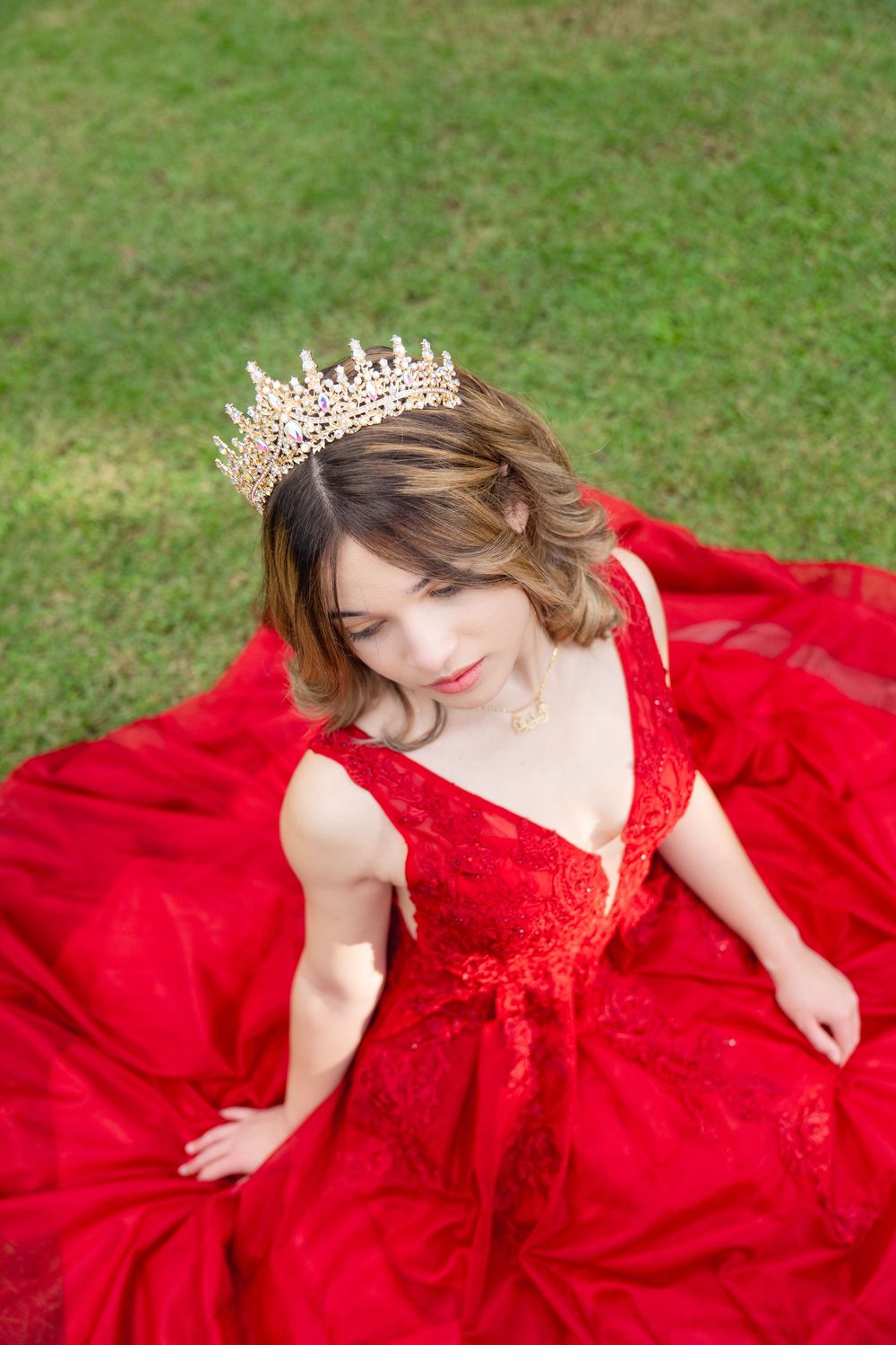 Young woman in red lace ball gown and gold crown sits gracefully on green grass, looking down elegantly.