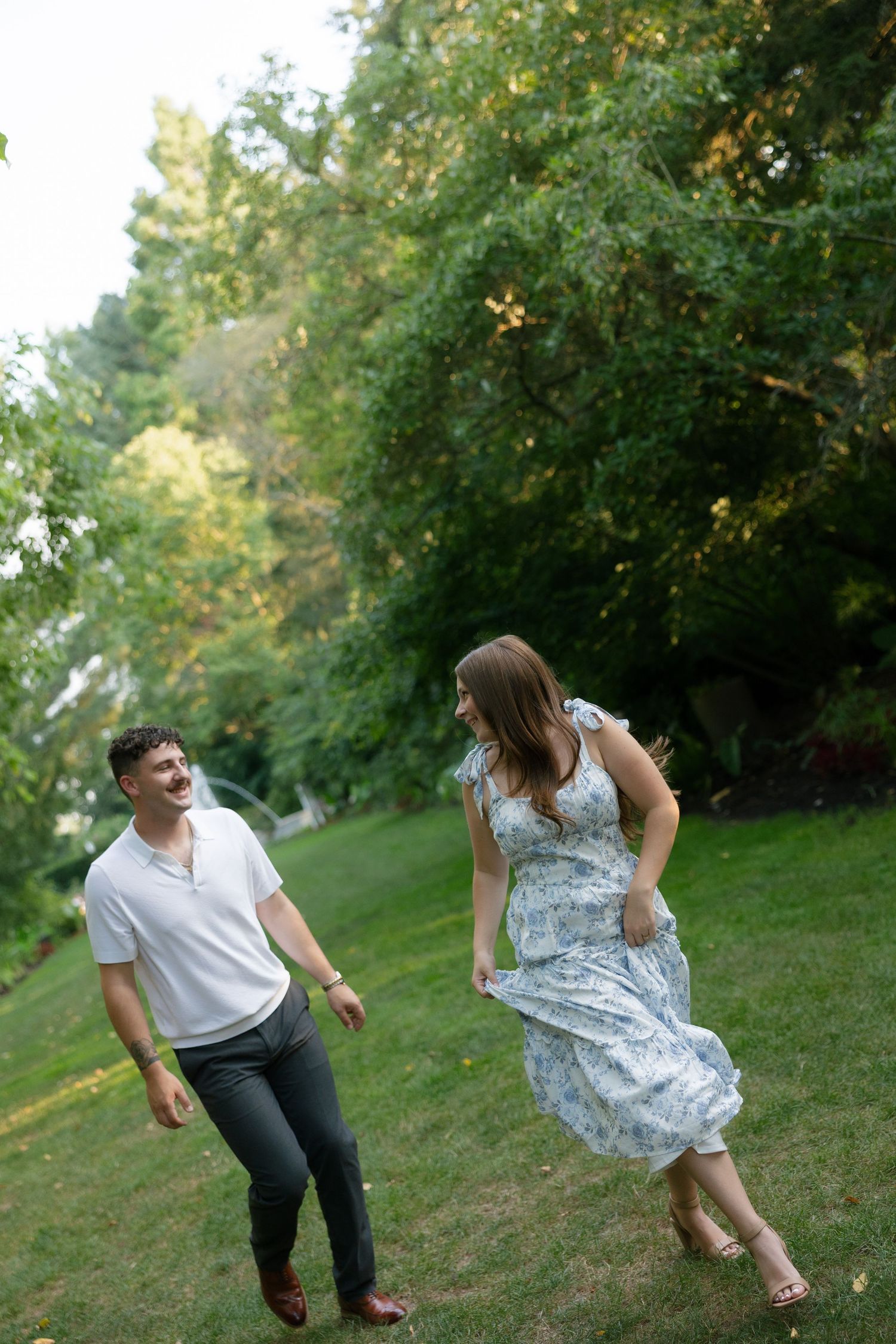 Laughing couple playfully walking together on a green lawn surrounded by tall trees.