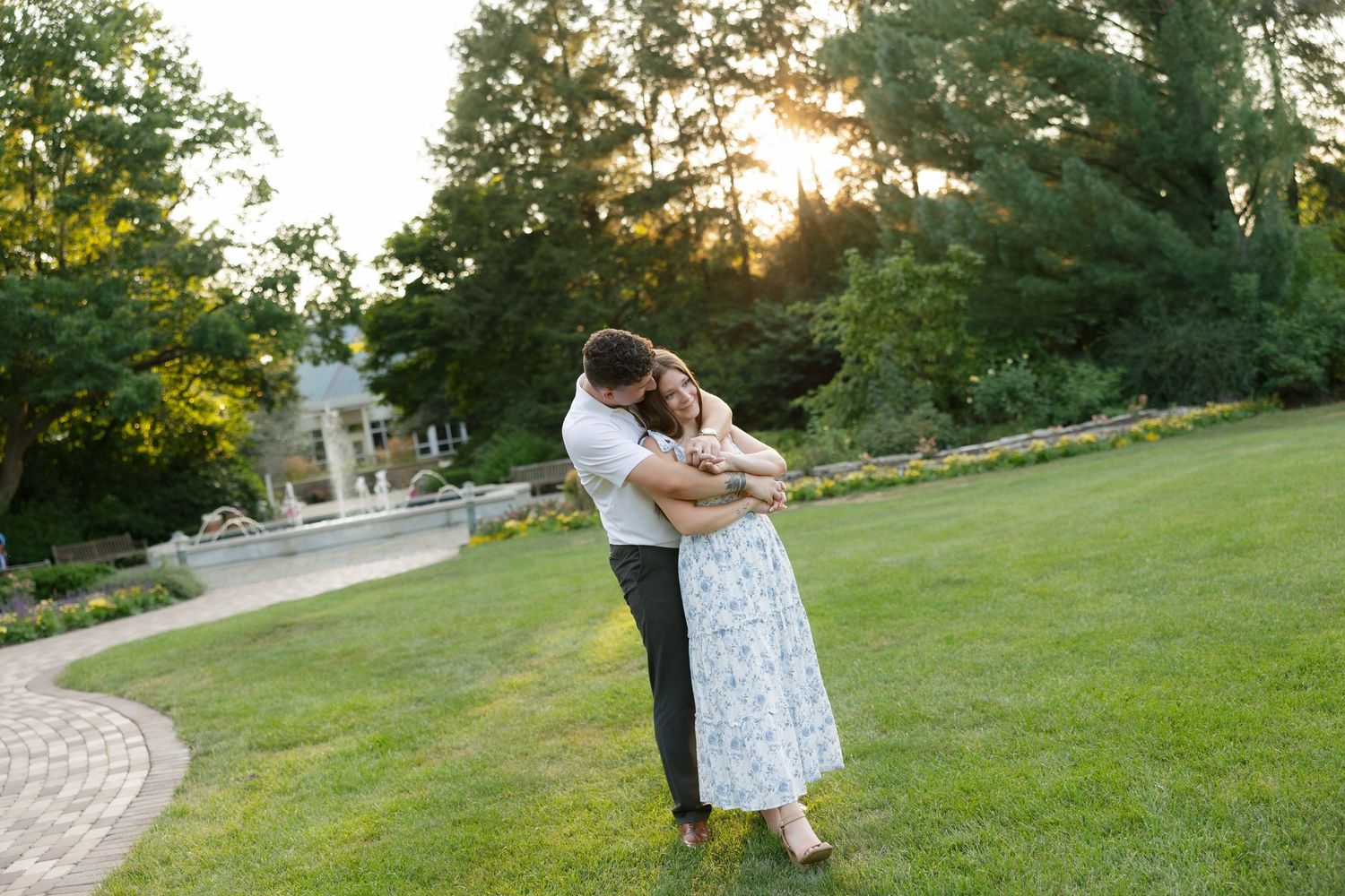Couple sharing a warm embrace on a garden lawn with a fountain and sunset backdrop.