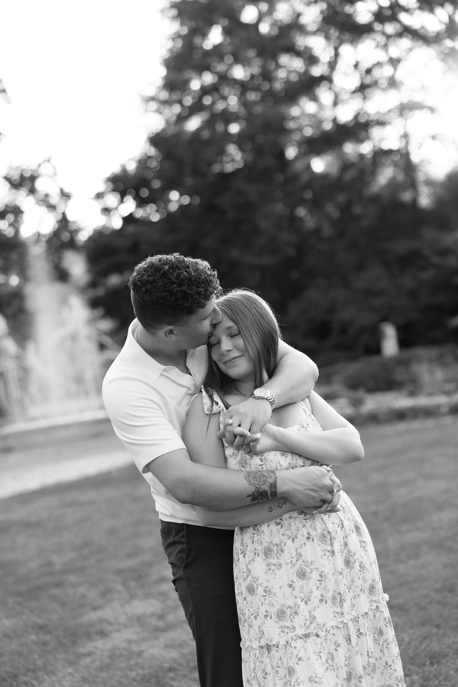 Black and white photo of a couple sharing a tender embrace in a park with a fountain.