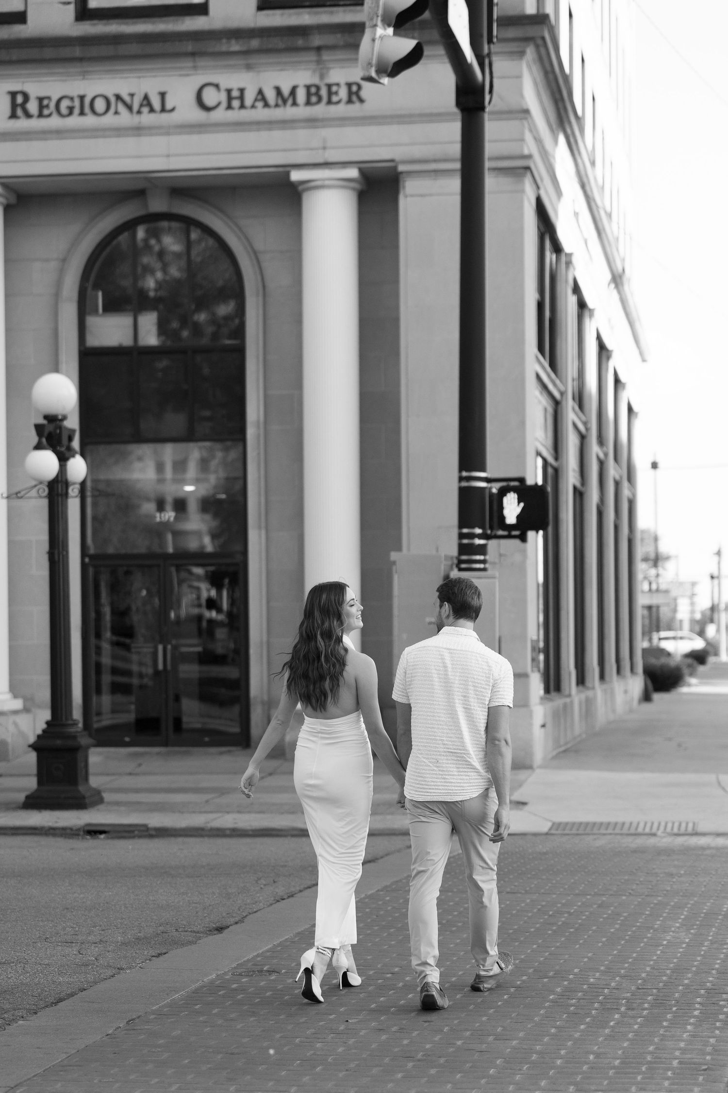 Black and white photo of couple walking hand in hand past The Regional Chamber building on a city street.