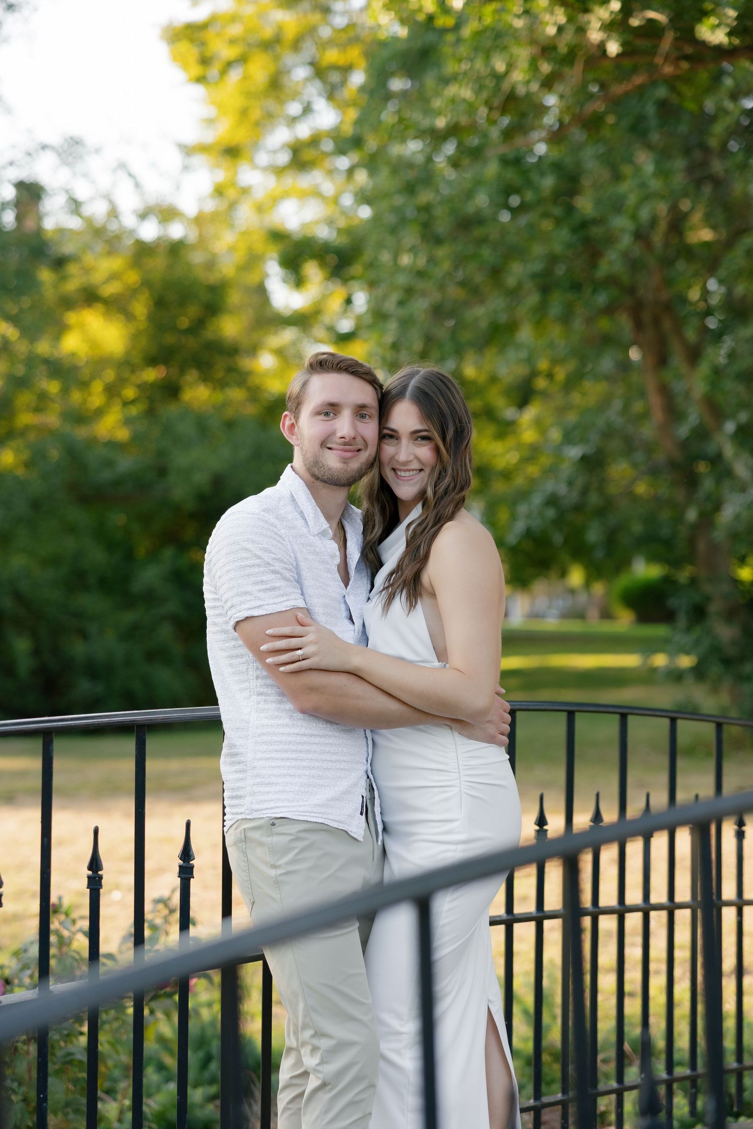 Happy couple embracing and smiling on an iron railing bridge surrounded by lush green trees.