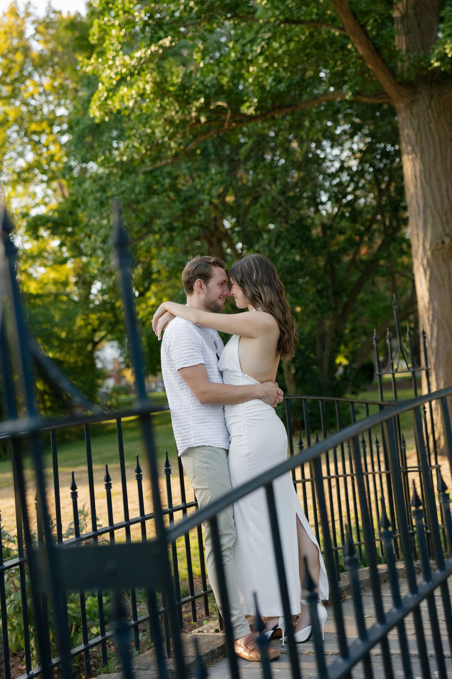 Romantic couple sharing a kiss on an ornate iron fence in a sunlit park setting.