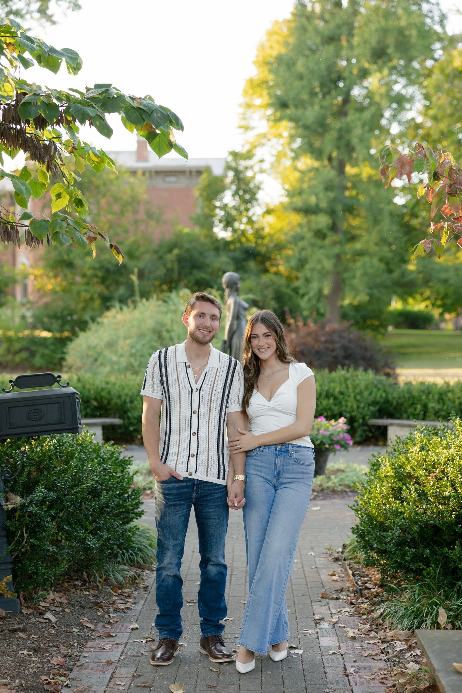 Smiling couple holding hands on a brick pathway surrounded by garden greenery and trees.