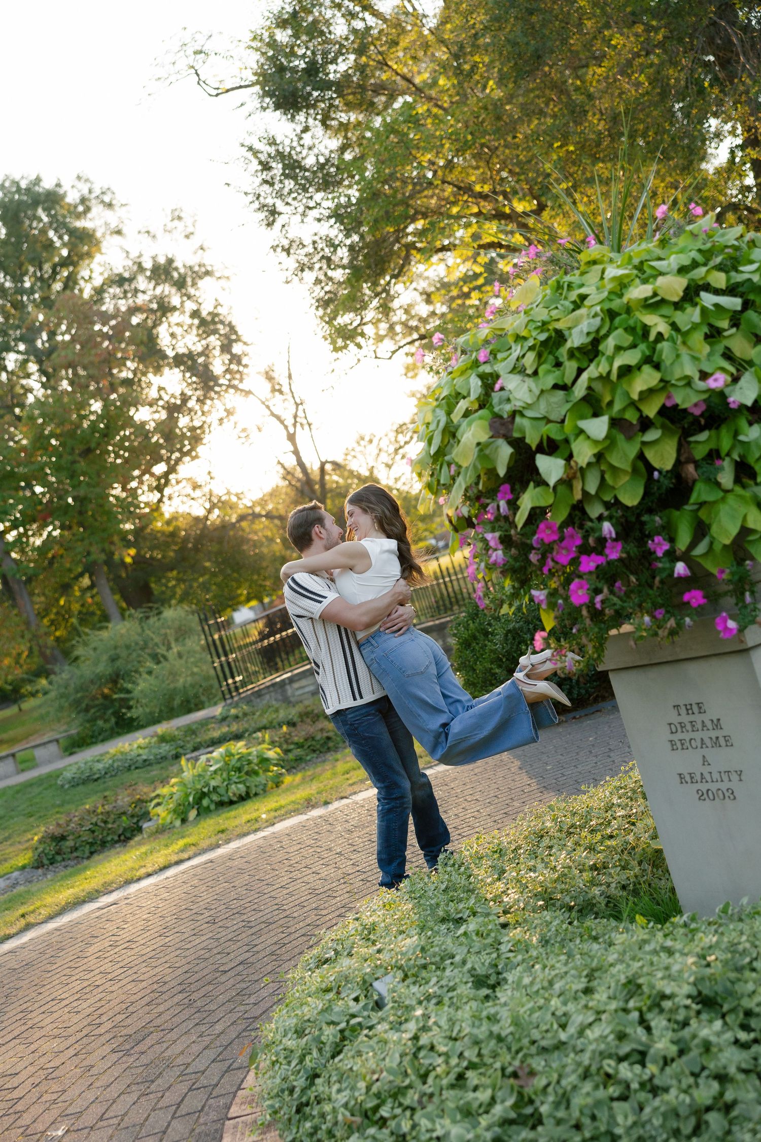 Man lifting woman in a romantic embrace on a brick path beside vibrant pink flowers.