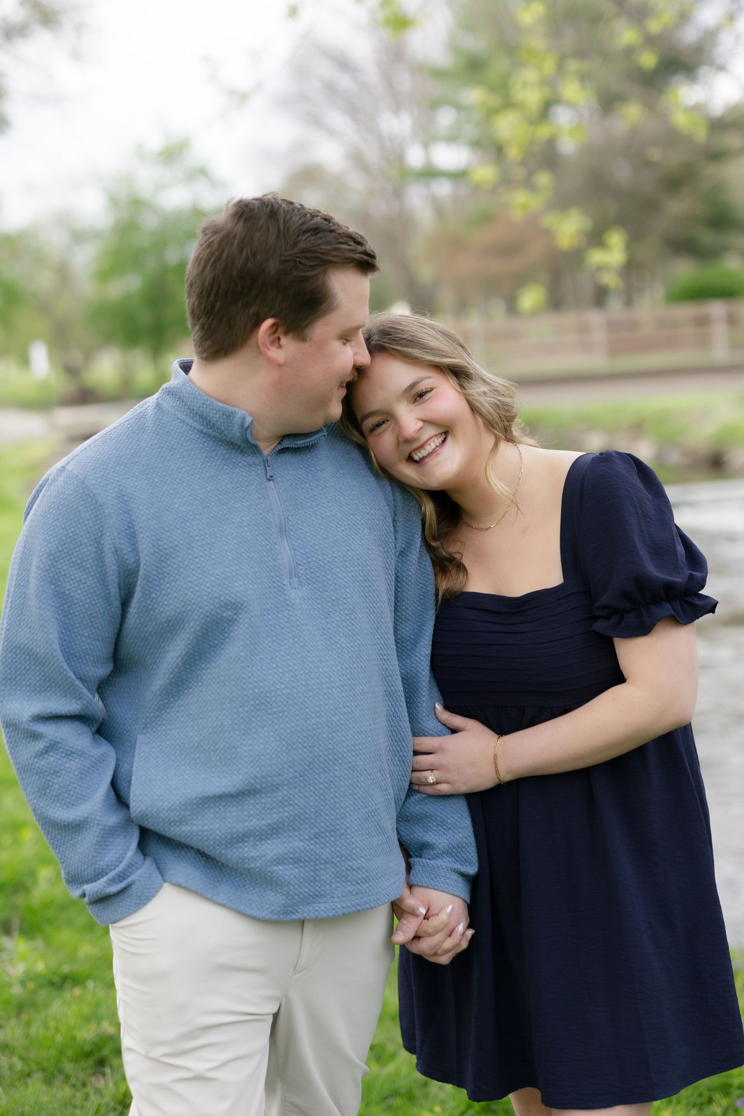 Smiling couple in a park, woman in navy dress leaning on man in blue pullover holding hands outdoors.
