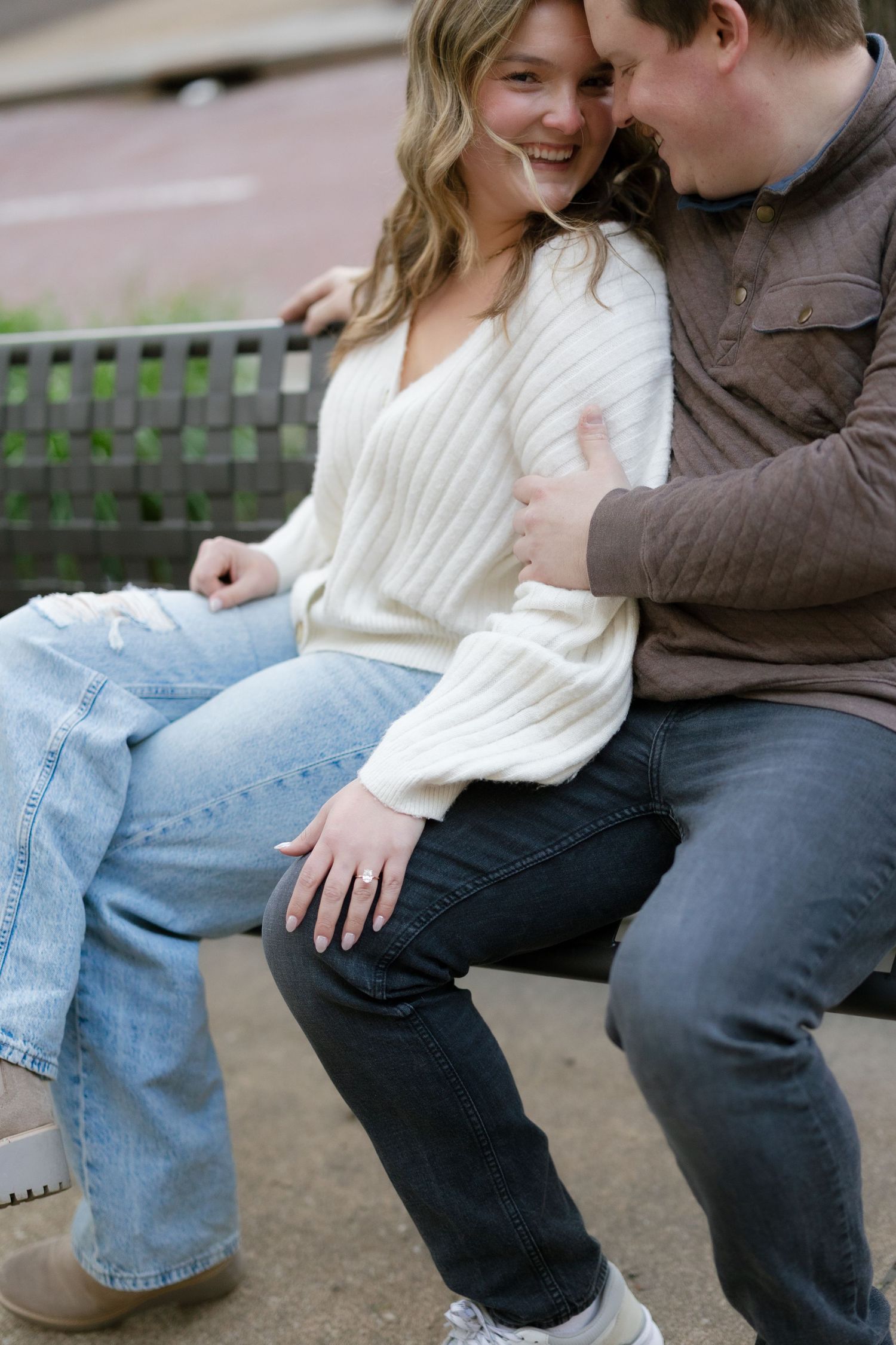 Happy couple sitting closely on a bench, woman in cream sweater and jeans laughing during engagement session.
