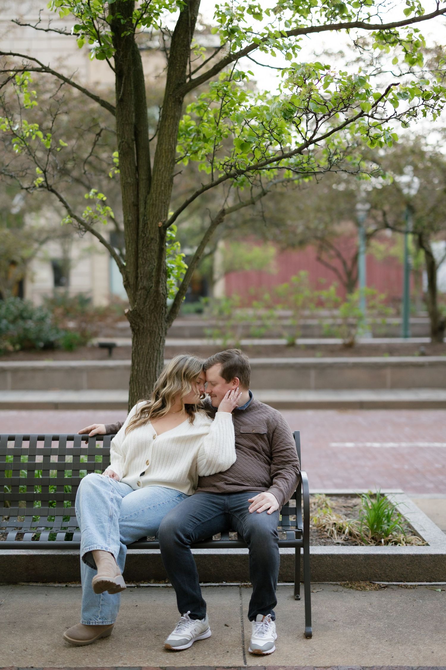 Couple sharing a tender kiss on a park bench surrounded by spring trees during outdoor engagement photos.