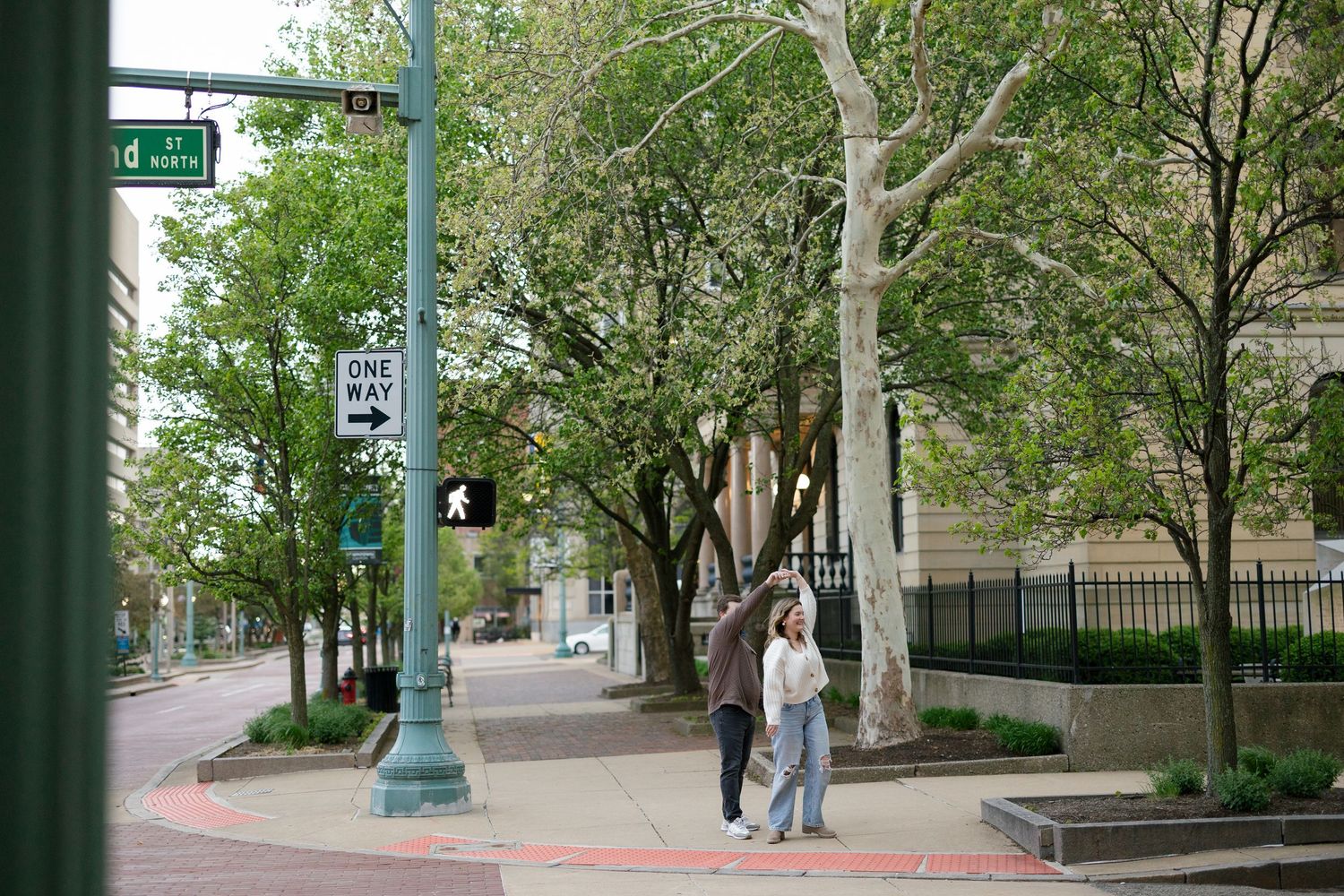 Couple walking together on a tree-lined urban sidewalk during a casual outdoor engagement photo session.