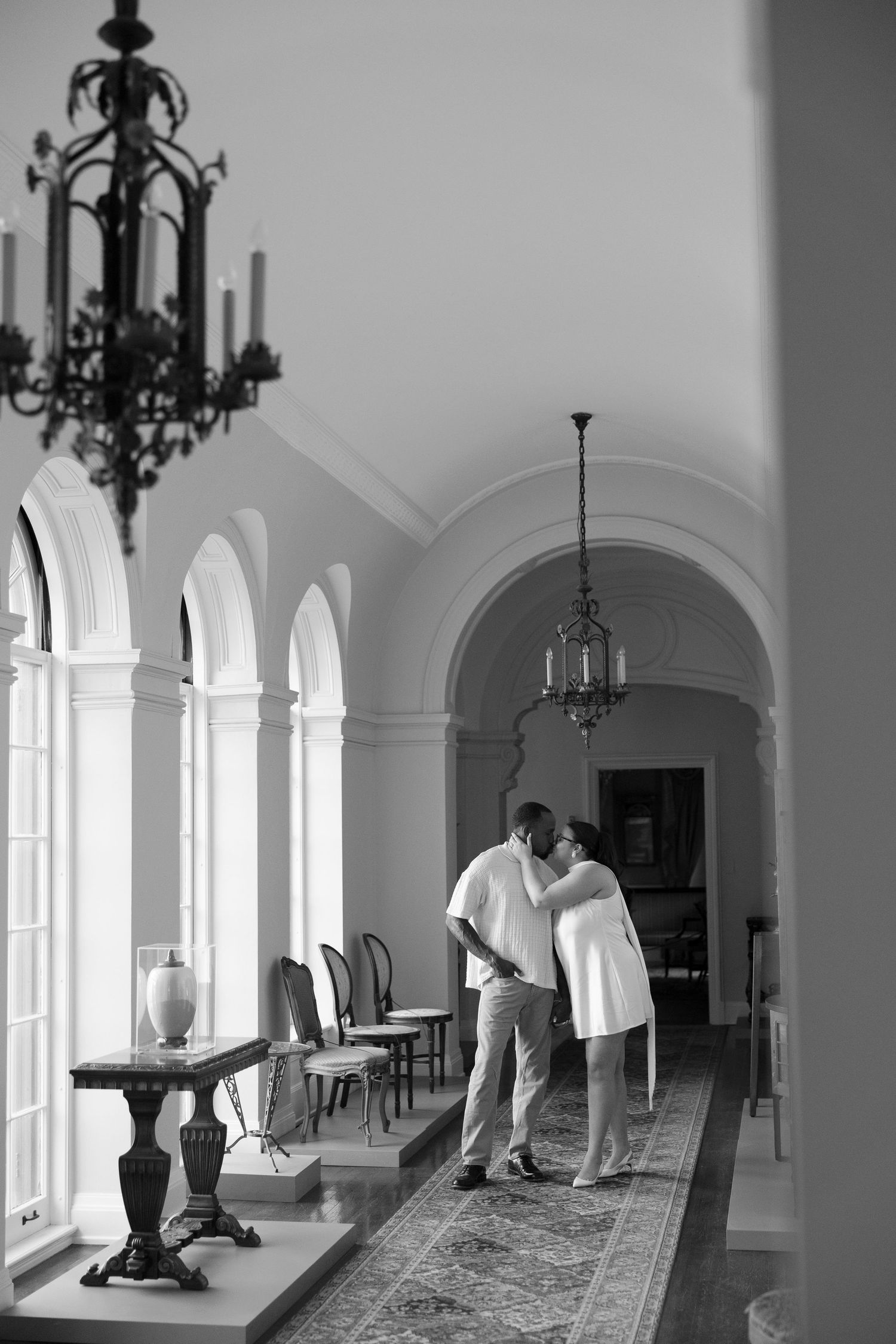 Black and white photo of couple embracing in an elegant arched hallway with chandeliers.