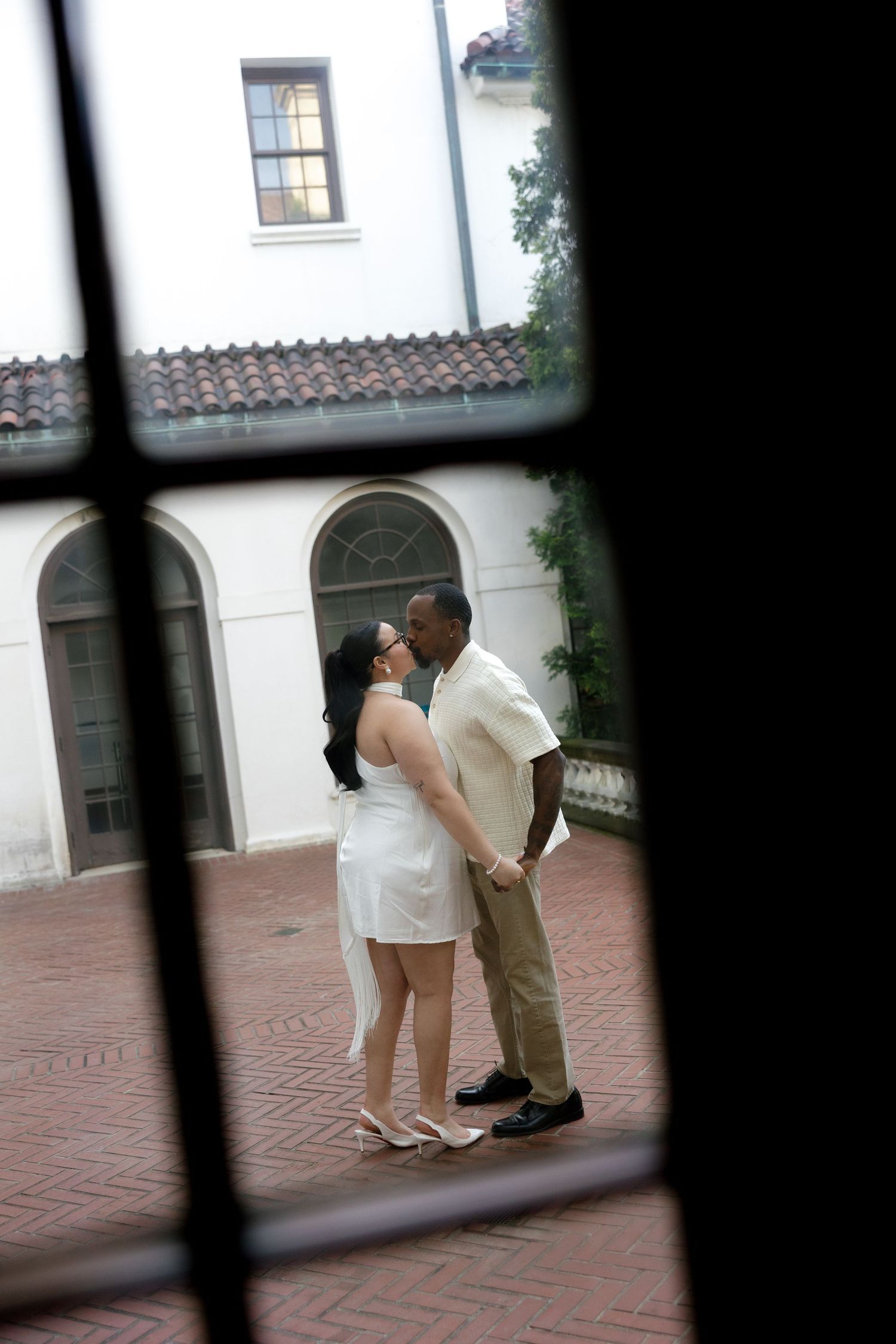 Couple sharing a kiss in a courtyard, photographed through a decorative window frame.