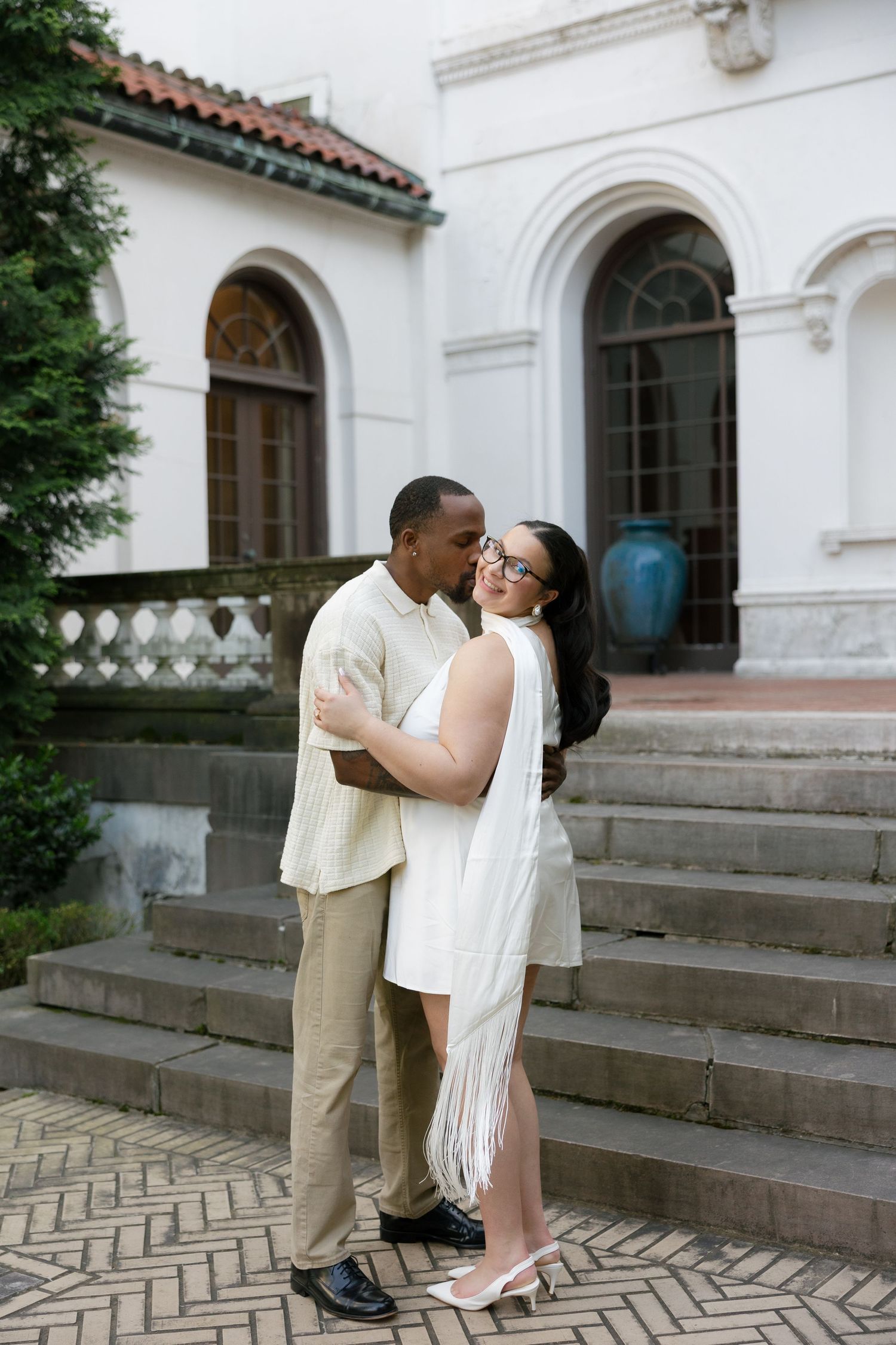 Interracial couple embracing on steps outside a white Mediterranean-style building with arched windows.