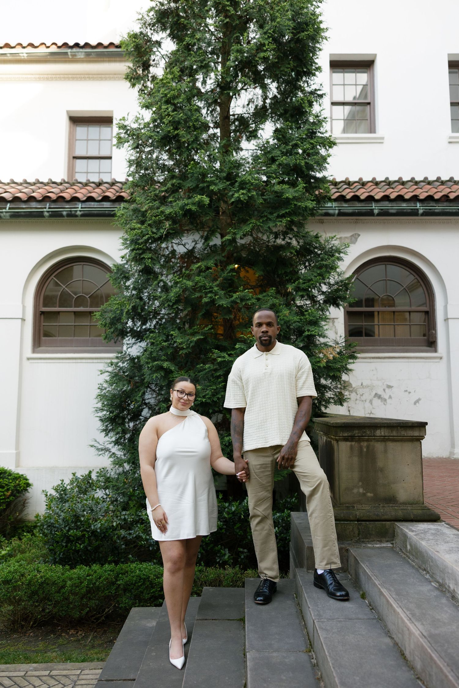 Couple holding hands in front of a white building with tall evergreen tree, woman in white dress.
