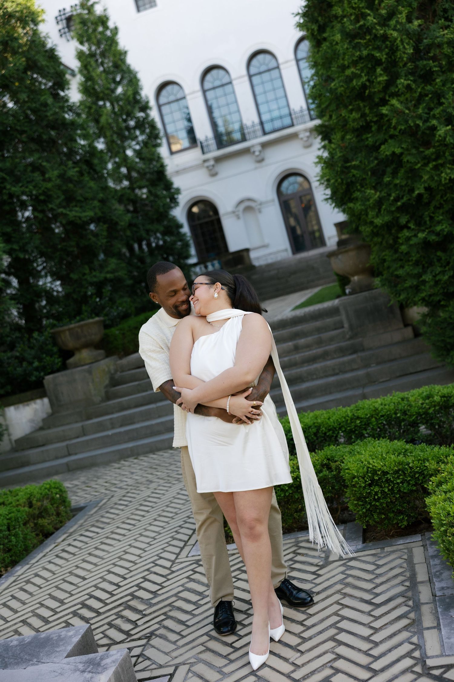 Interracial couple embracing on herringbone stone steps outside a grand white building surrounded by greenery.