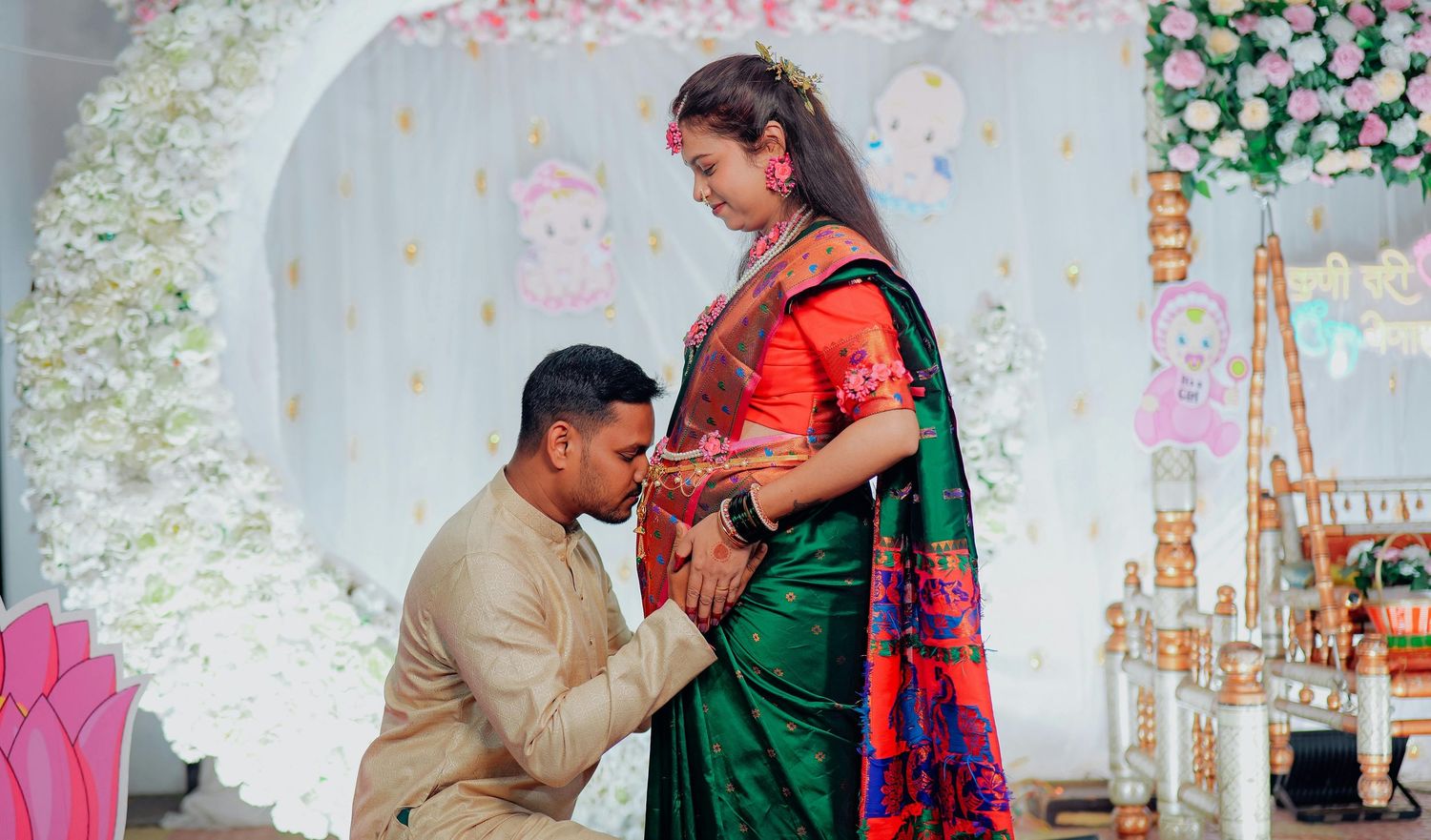 Man in beige kurta kissing pregnant woman's baby bump during Indian baby shower ceremony with floral decor.