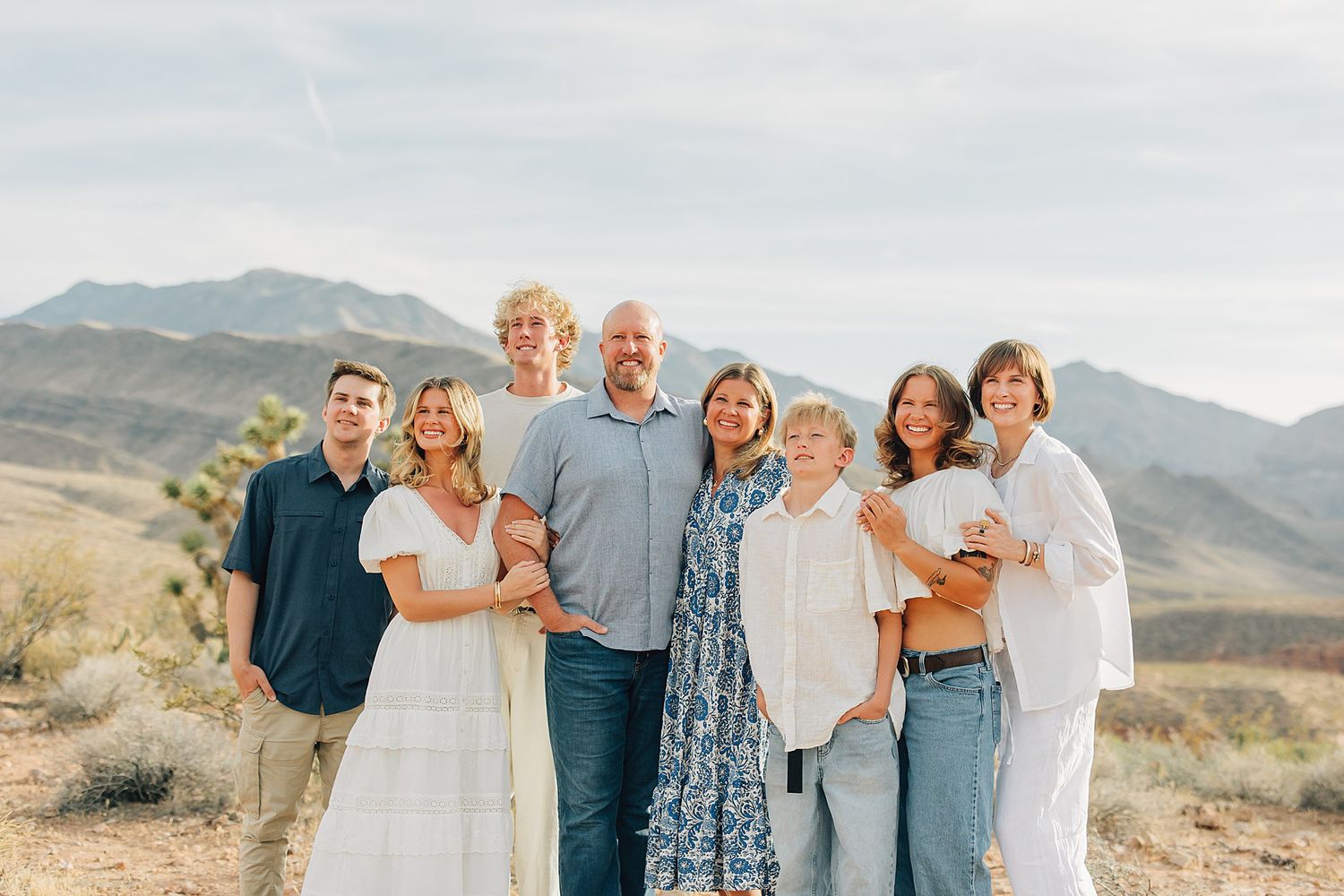 Family of eight smiling together in desert landscape with mountains and Joshua trees in background.