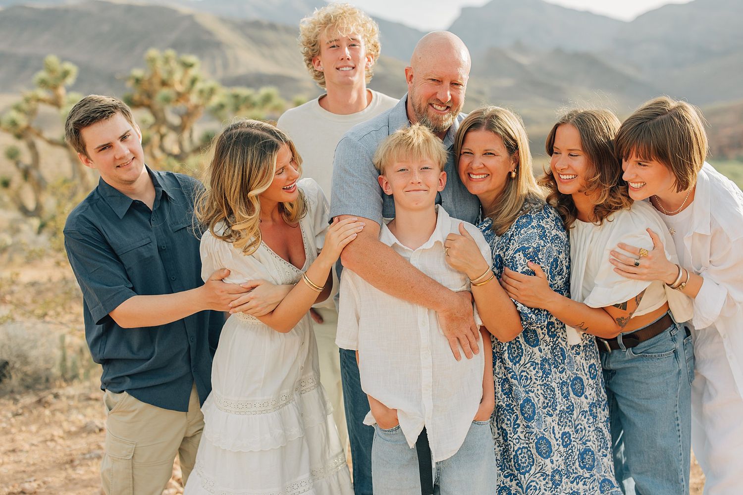 A joyful family of eight poses together outdoors in a desert landscape with Joshua trees and mountains in the background.
