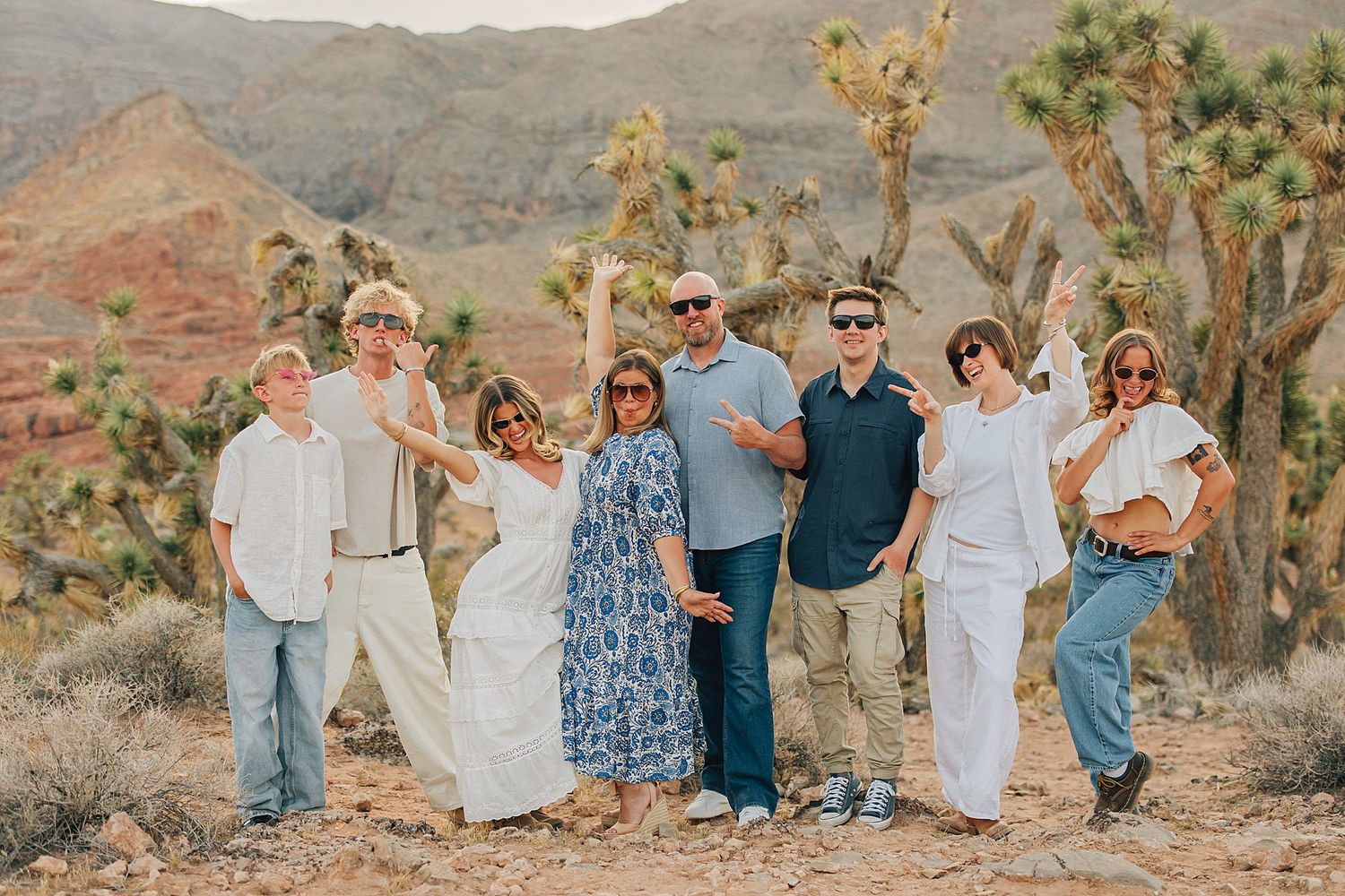 Family of seven posing happily in Joshua Tree desert landscape with mountains and cacti backdrop.