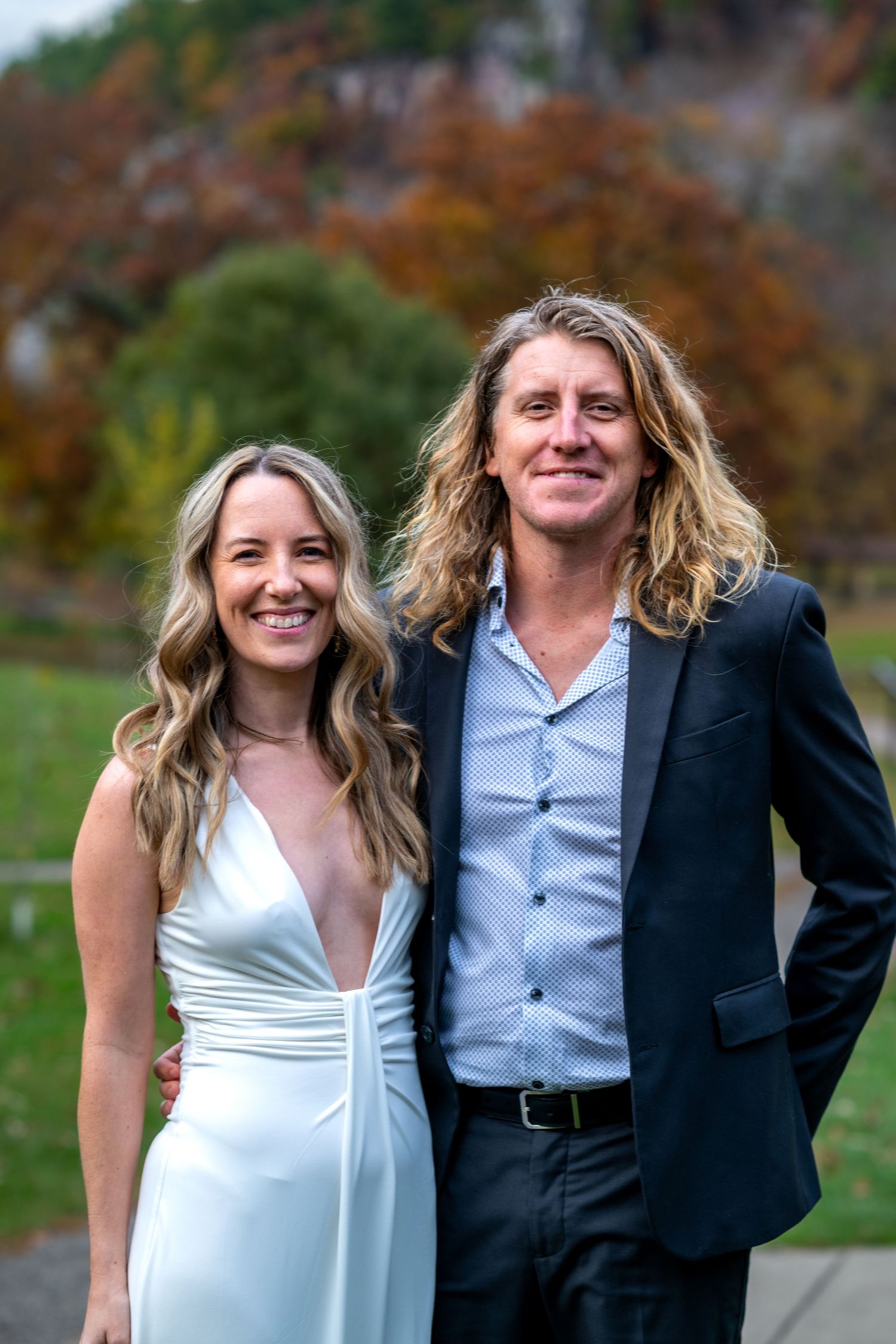 A smiling couple in formal attire pose outdoors in autumn, woman in white gown and man in dark suit with long hair.