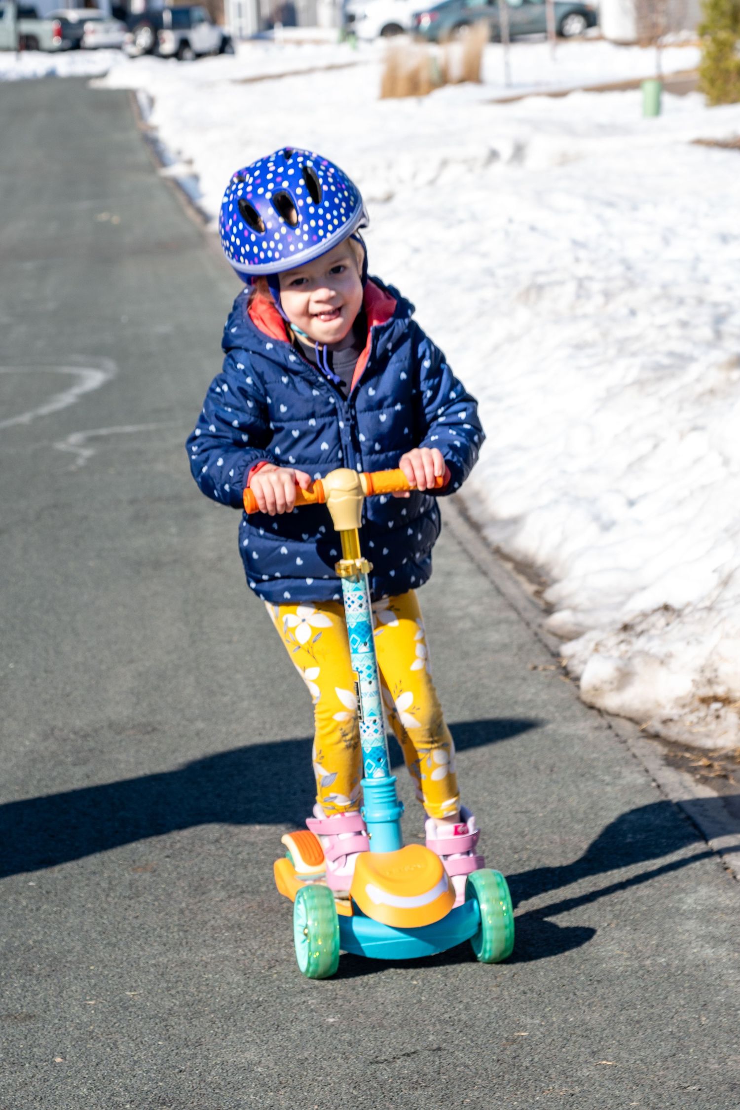 Young girl in blue polka dot jacket and helmet rides colorful three-wheel scooter on sunny winter day.