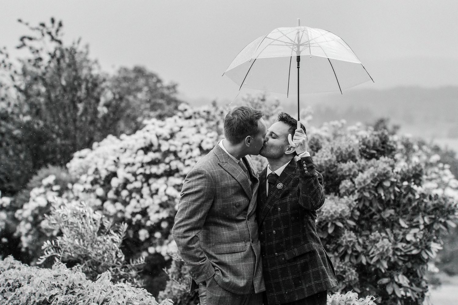 Two grooms in suits, just married, holding an umbrella in the rain and kissing