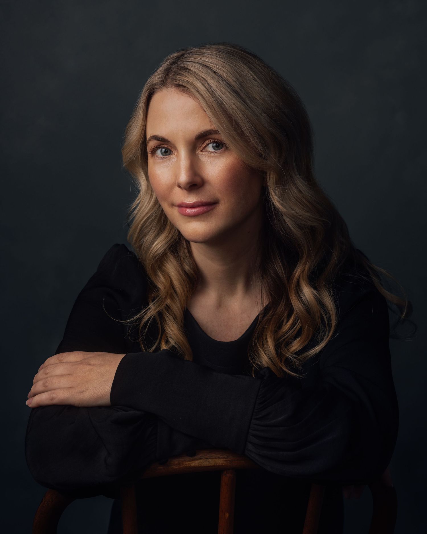 A blonde woman in a black outfit poses elegantly against a dark studio background, arms crossed on a wooden stool.