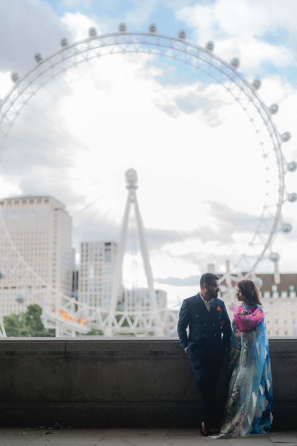 Couple in elegant attire posing in front of the London Eye Ferris wheel on a cloudy day.