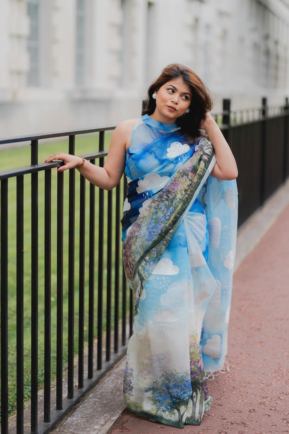 Woman in blue sky-print saree poses gracefully by black iron railing outdoors near a white building.